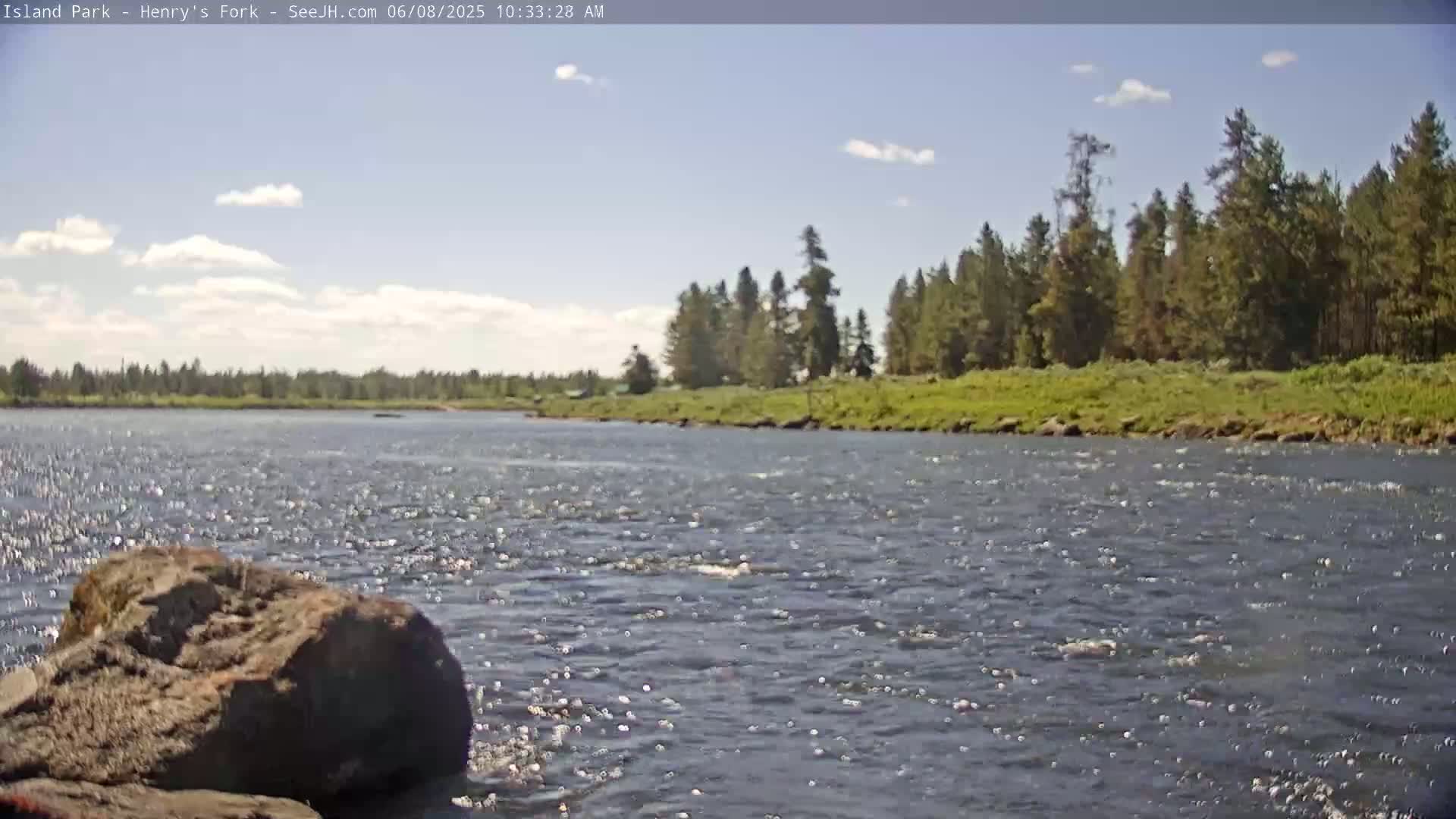 A mostly sunny day shows a river flowing past a rocky shoreline and a forested bank under a partly cloudy sky.