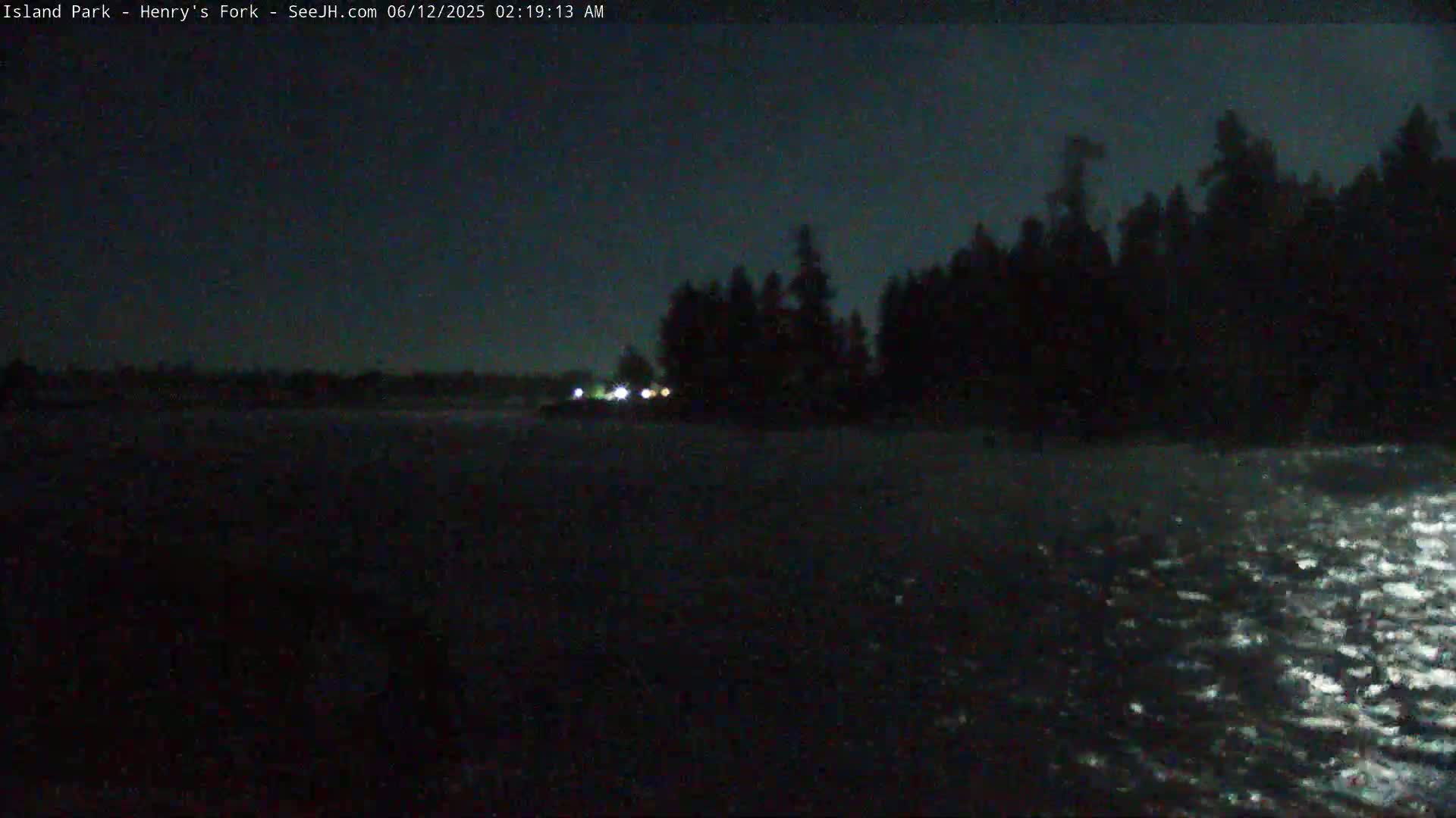 A dark, nighttime scene shows a body of water reflecting light, with a silhouetted treeline visible in the distance under a mostly clear sky.