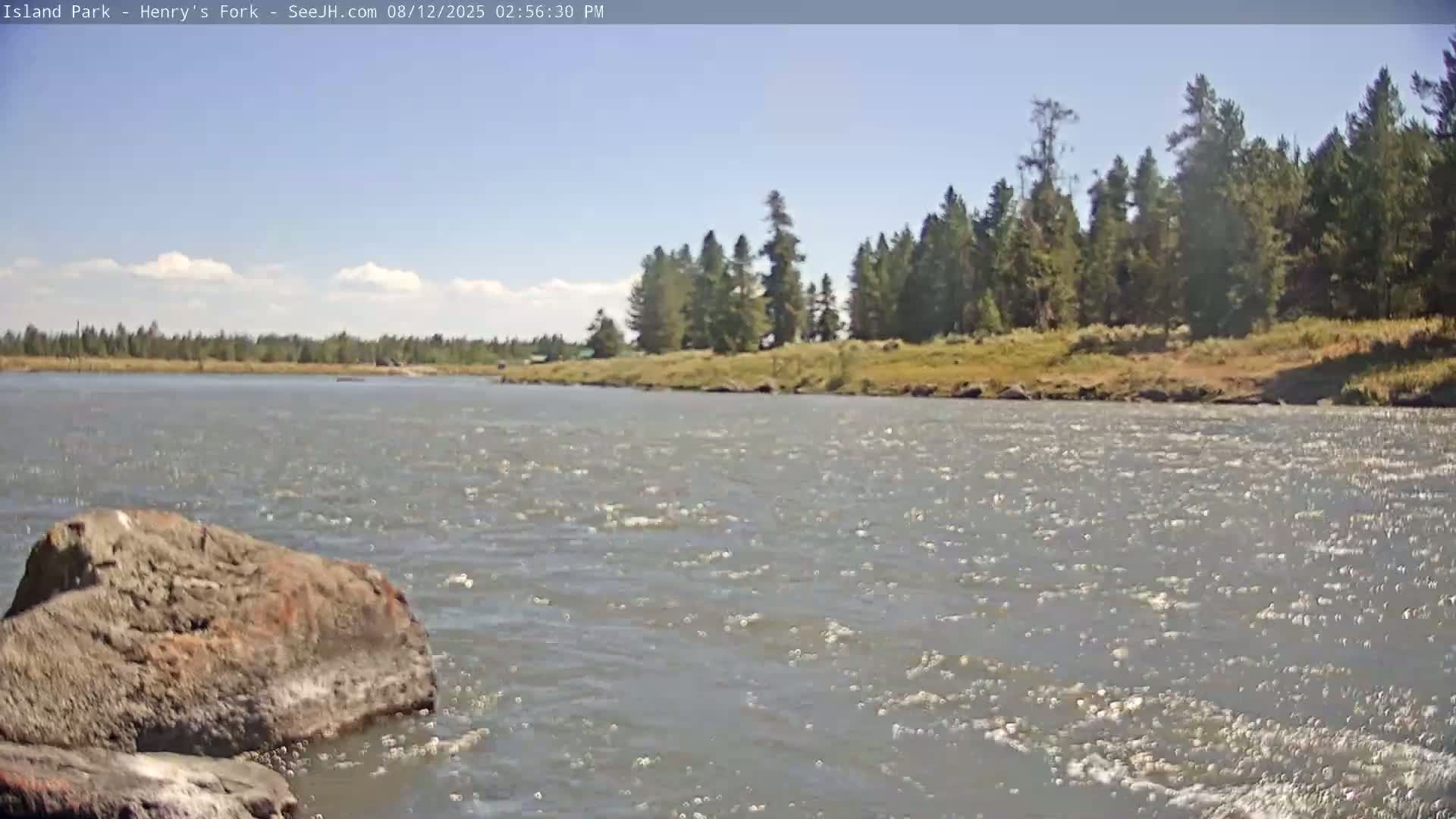 A rocky river flows under a partly cloudy sky past a grassy bank lined with evergreen trees.
