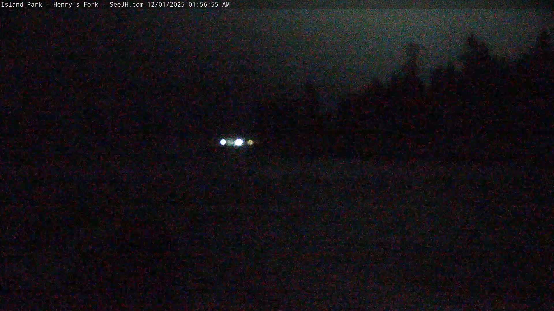 A very dark and grainy outdoor nighttime scene under an overcast sky features three bright, distant lights with a faint treeline visible on the right.