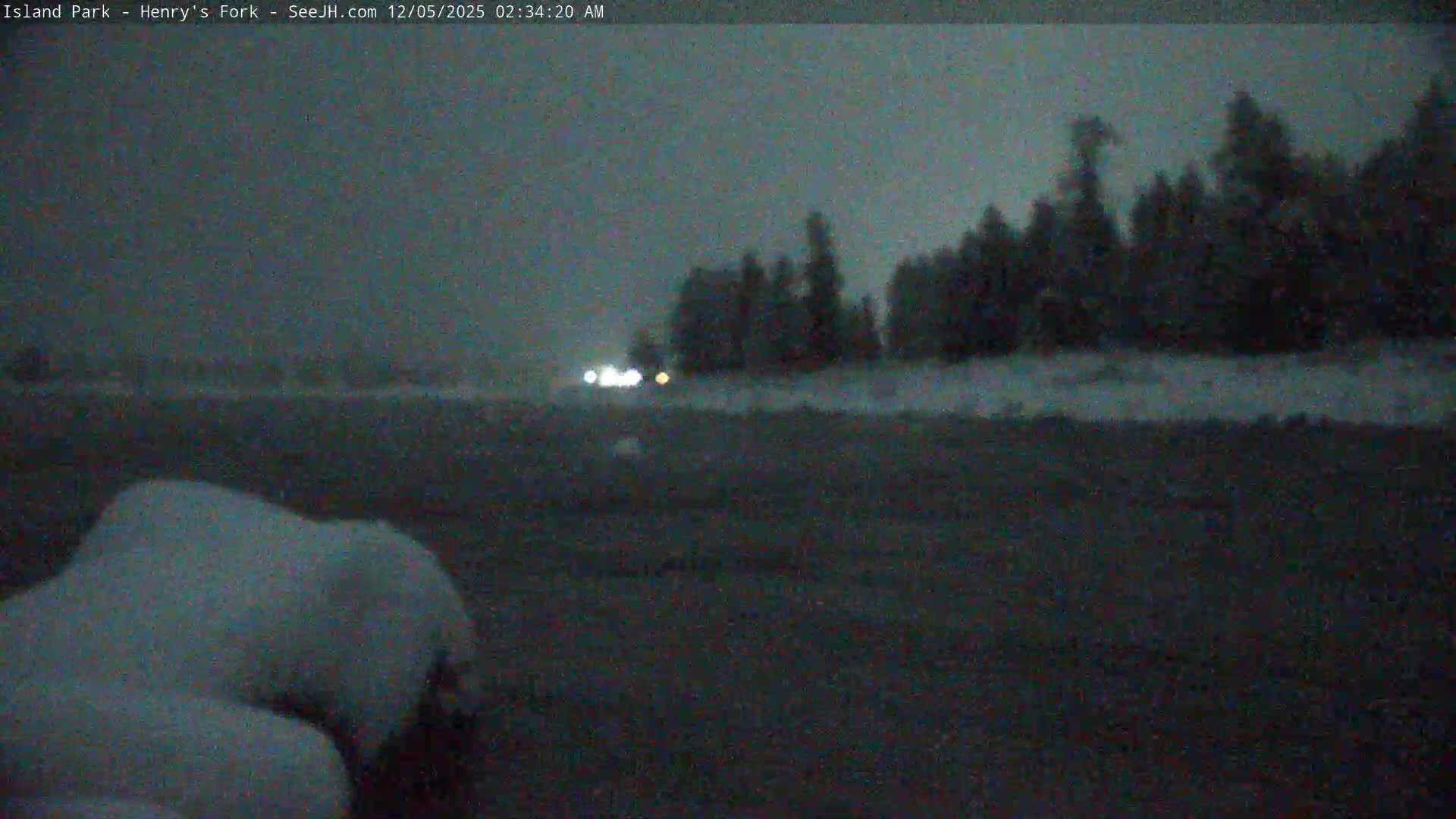 A dark, hazy winter night scene reveals a prominent snowdrift in the foreground, with faint distant vehicle lights and silhouetted trees under an overcast sky with low visibility.