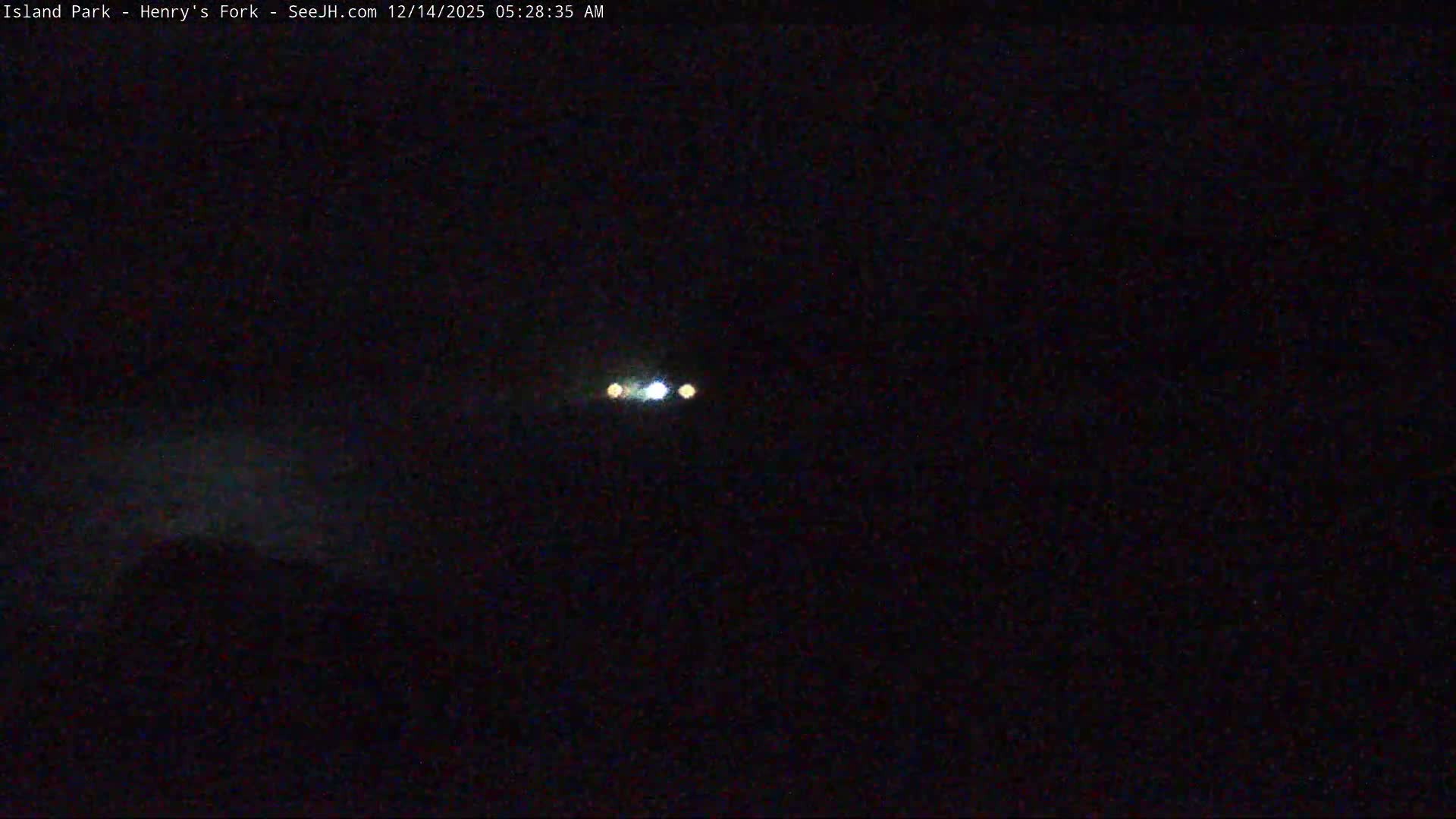 A dark, hazy winter night scene reveals a prominent snowdrift in the foreground, with faint distant vehicle lights and silhouetted trees under an overcast sky with low visibility.