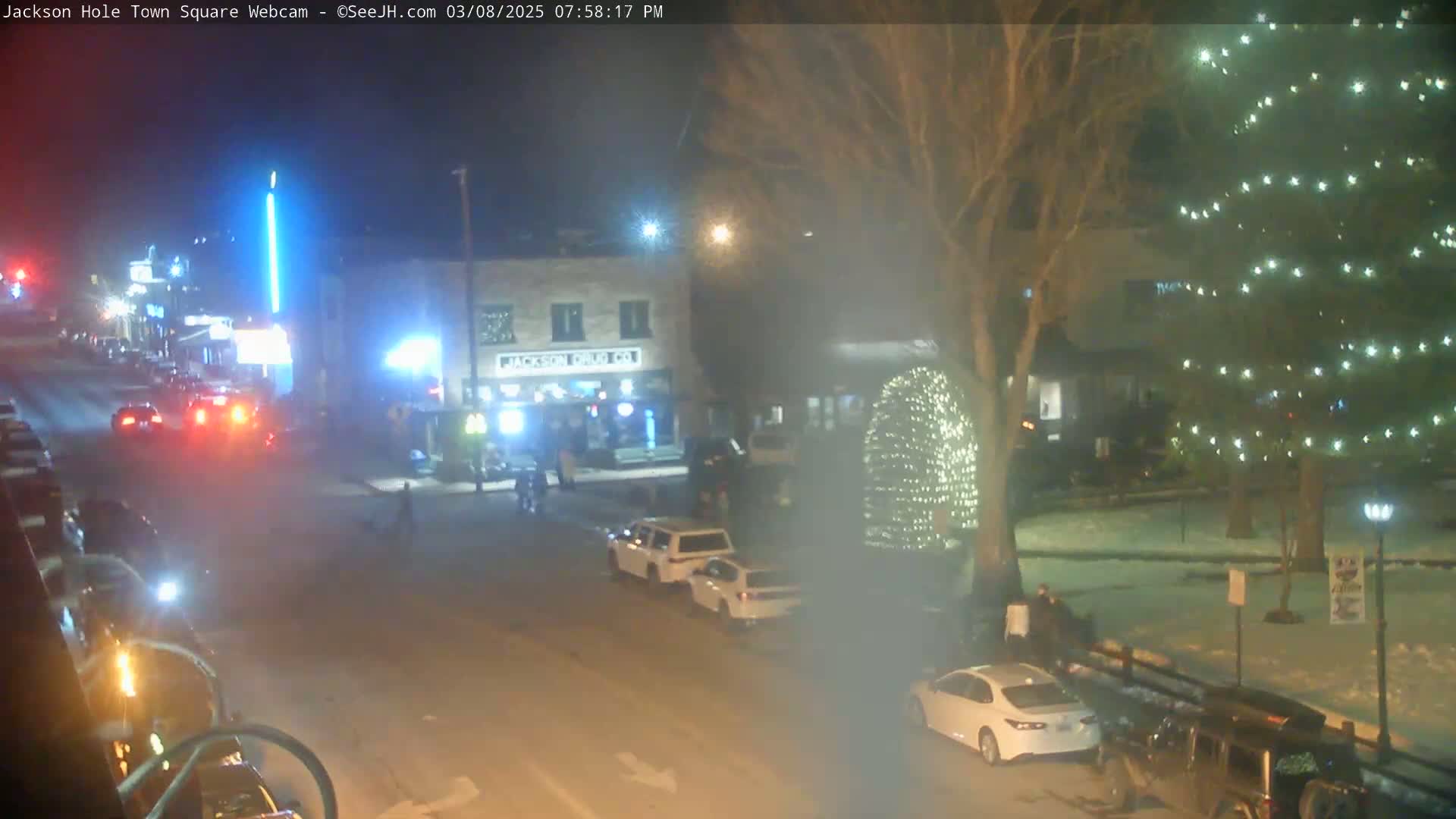 A nighttime street scene shows cars parked along a road, a few people walking, and illuminated Christmas trees and light displays in a snowy town square.