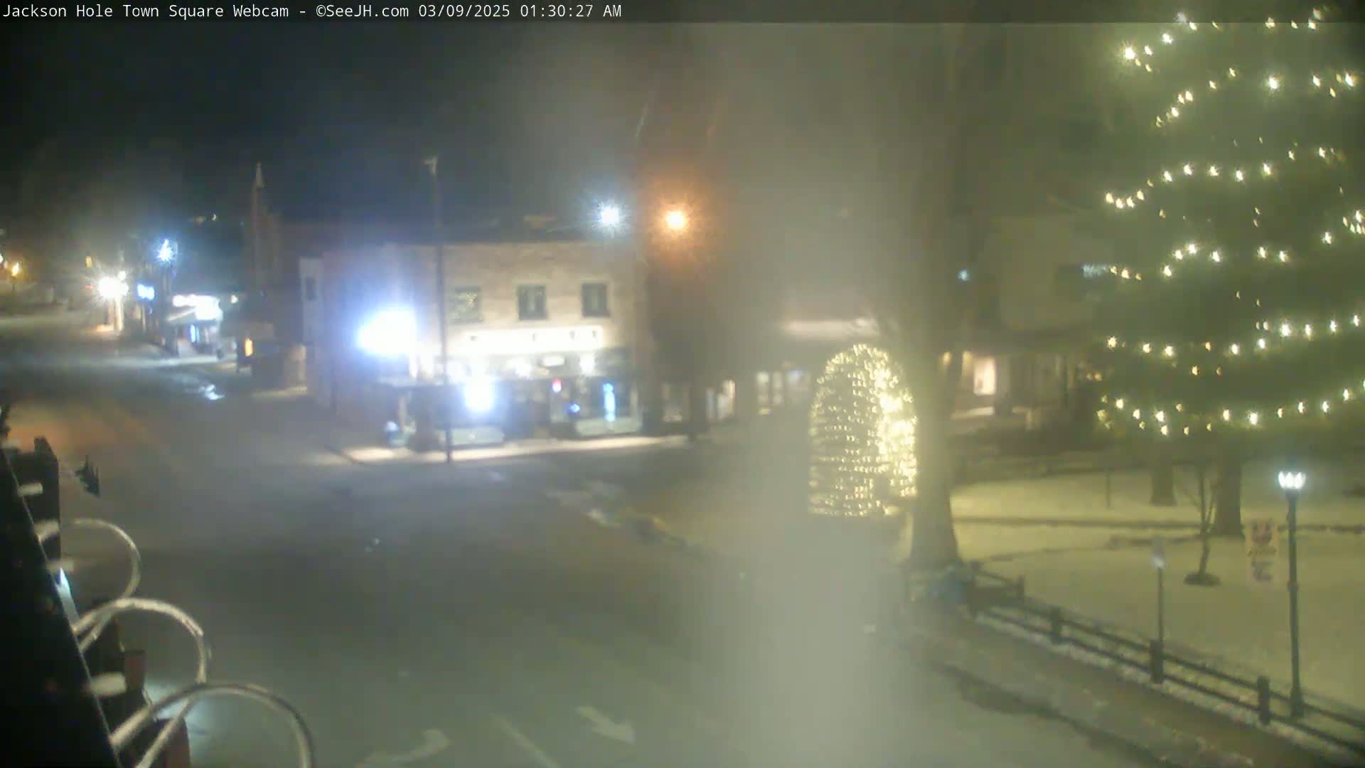 A nighttime view of a town square with some snow on the ground, illuminated by streetlights and Christmas lights on trees and other structures.