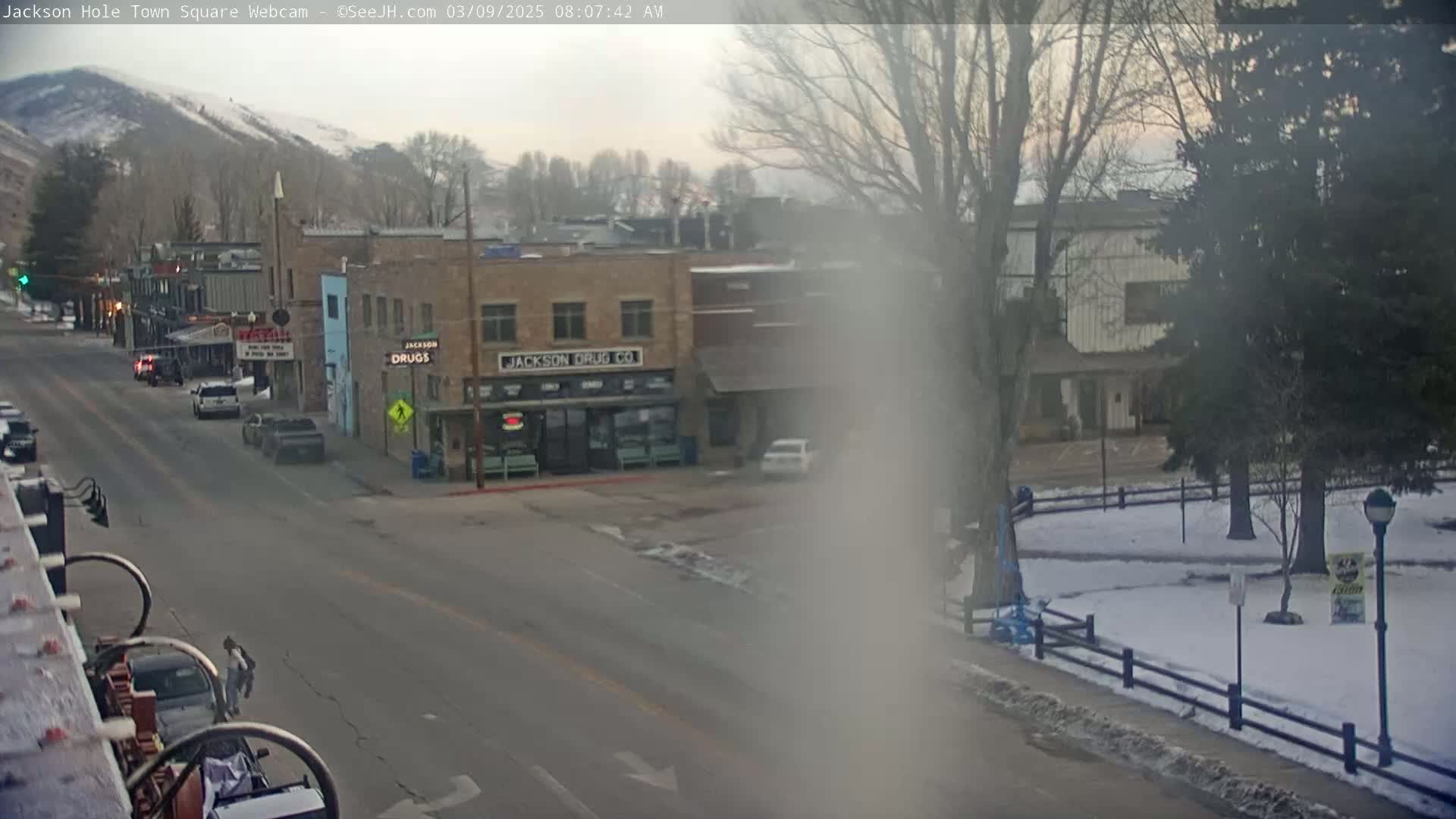 A mostly clear, cool morning view of a town square with snow on the ground in a park-like area and a few cars on the street, with mountains in the background.