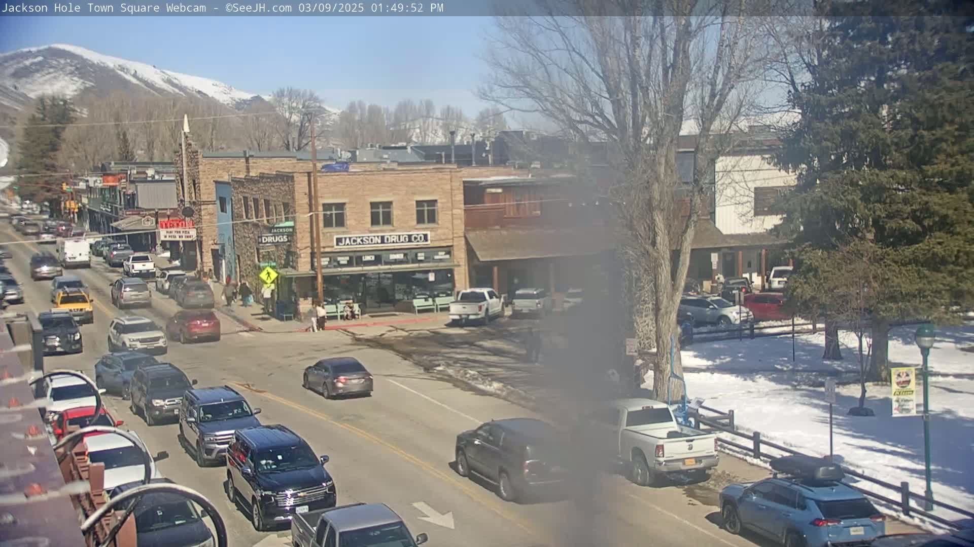 A sunny, snow-dusted town square scene shows several vehicles driving along a street lined with buildings, some with visible snow on the ground and nearby mountains in the background.