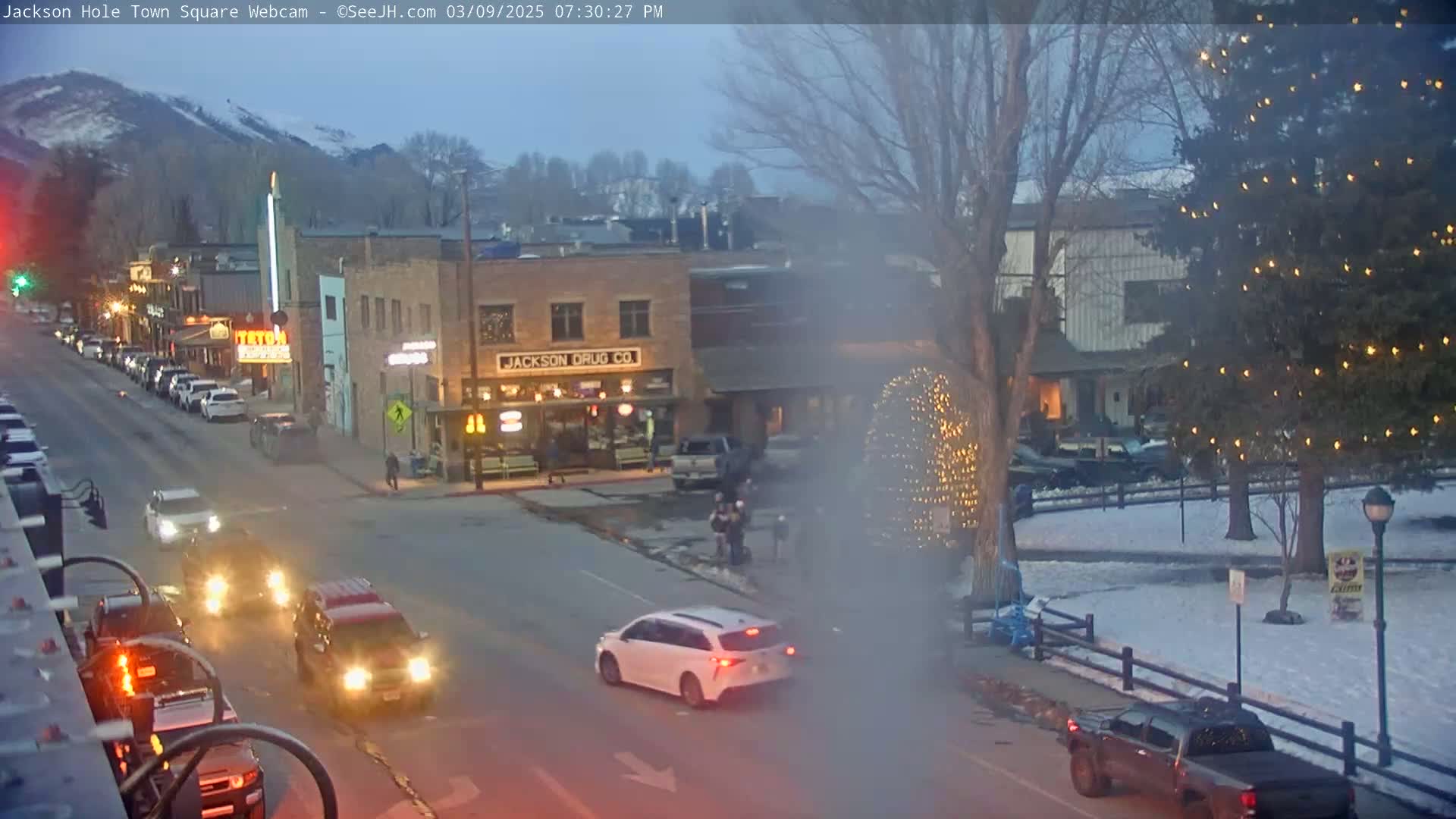 A town square at dusk, lightly snowing, with cars driving on a street past buildings and a decorated Christmas tree in a park.