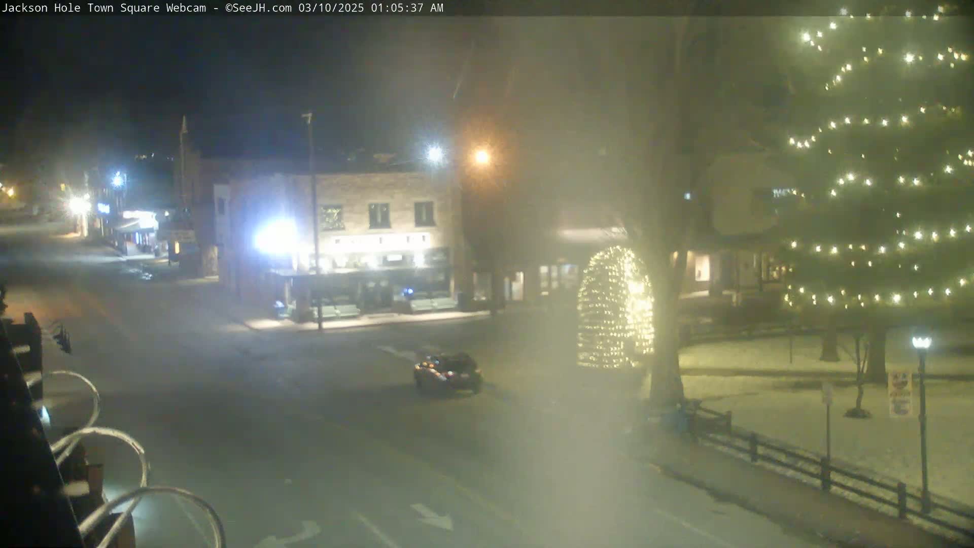 A nighttime view of a town square with a few buildings, a vehicle, and some illuminated trees in the snow.