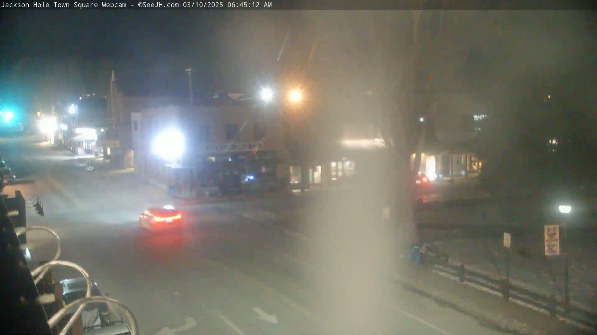 A mostly empty street scene at night in foggy conditions shows a few cars and buildings with lights on.