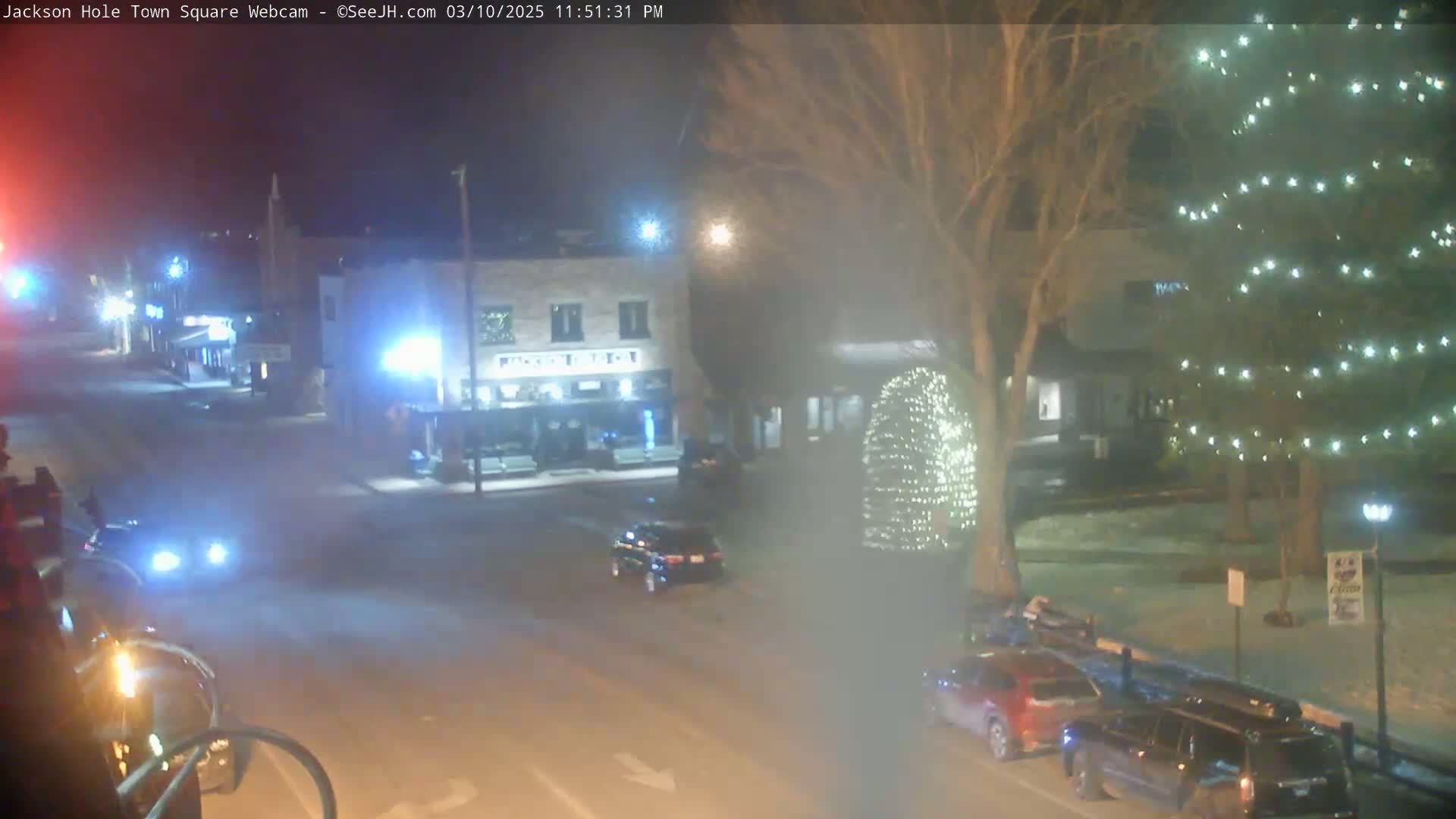 A nighttime view of a town square with cars on the street, snow on the ground, and a decorated tree.