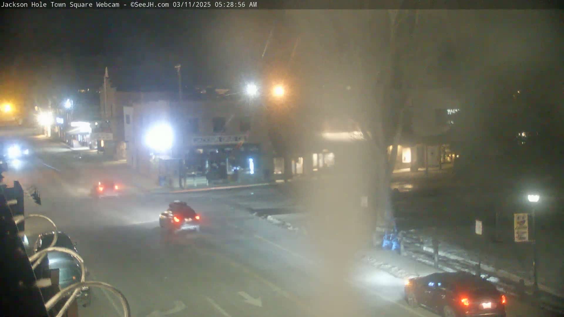 A nighttime, hazy view of a town square with a few cars on the road and streetlights illuminating the scene.