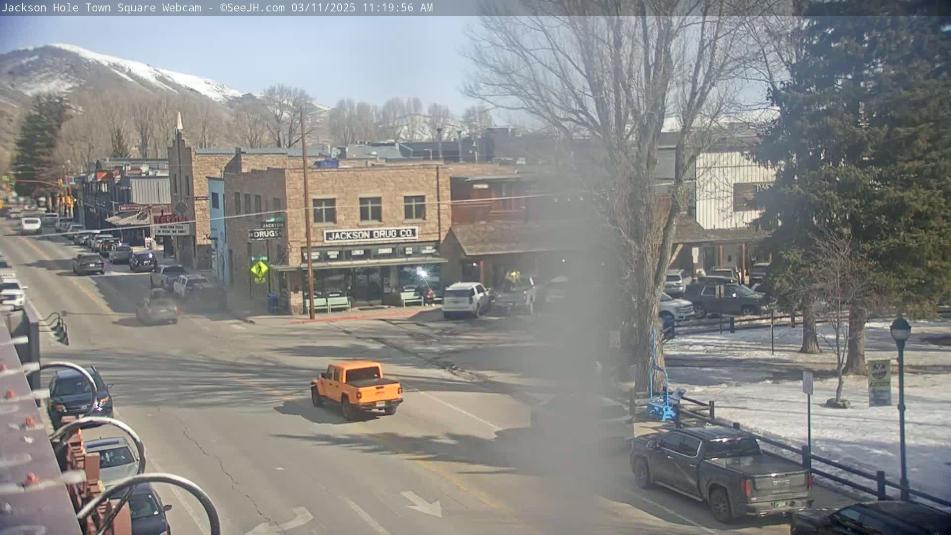 A partly sunny day reveals a town square with several vehicles, including a bright orange pickup truck, driving past brick buildings and trees, with a snow-capped mountain in the background.