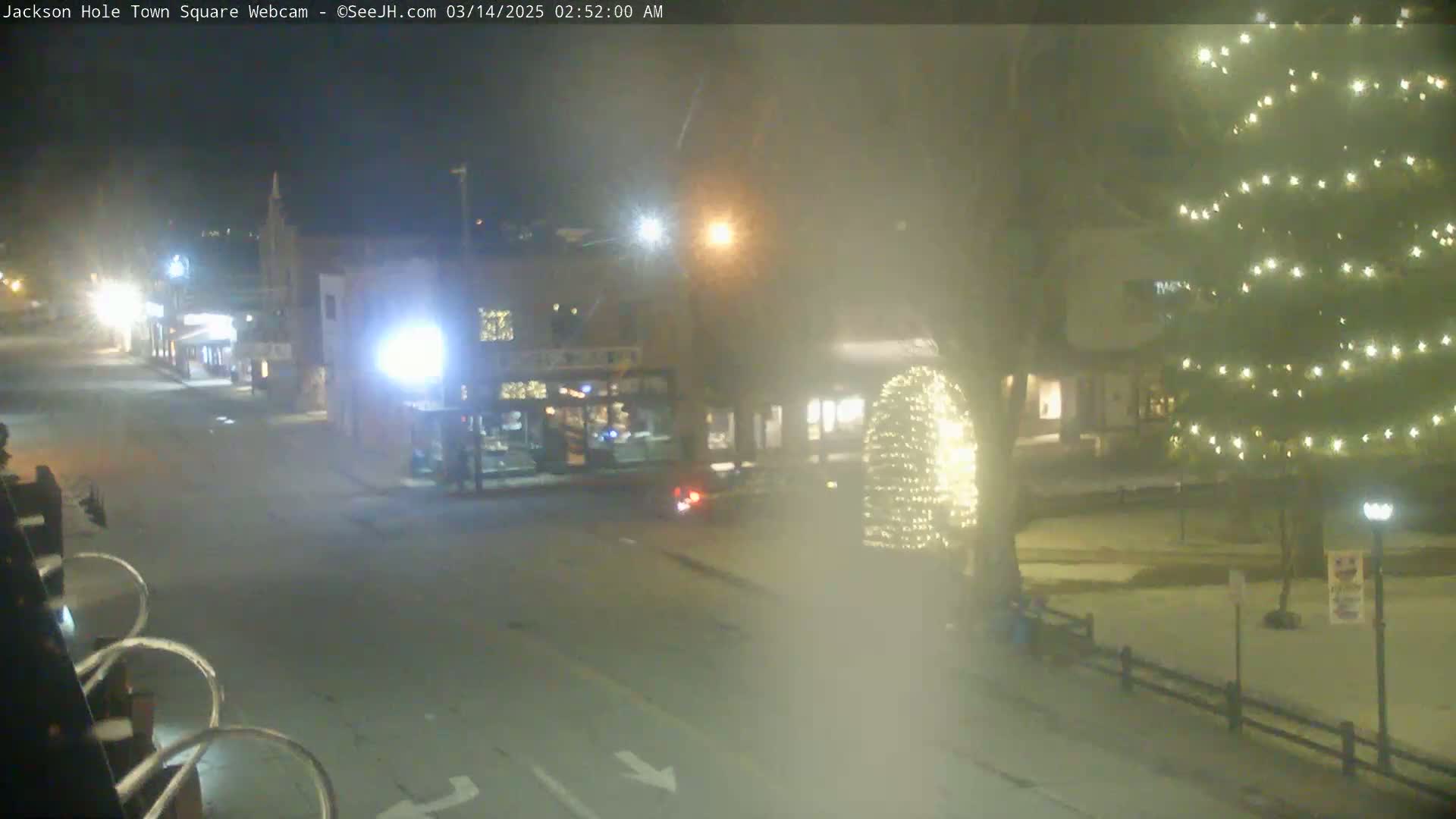 A nighttime view of a town square with snow on the ground, featuring a few illuminated buildings and a Christmas tree adorned with lights, all under hazy conditions.