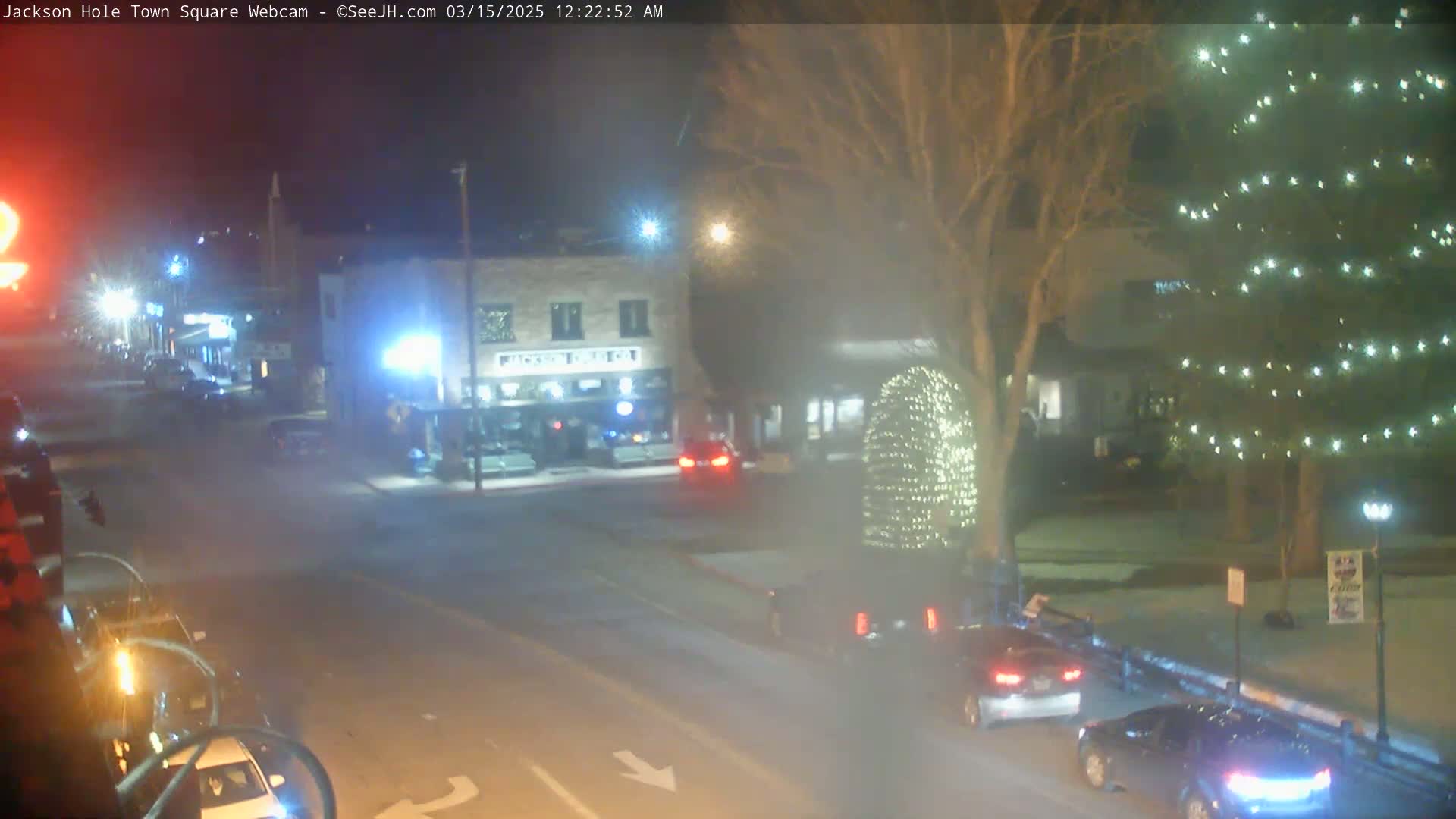 A nighttime, snow-dusted town square scene shows several cars at a road intersection near buildings and a tree decorated with lights.