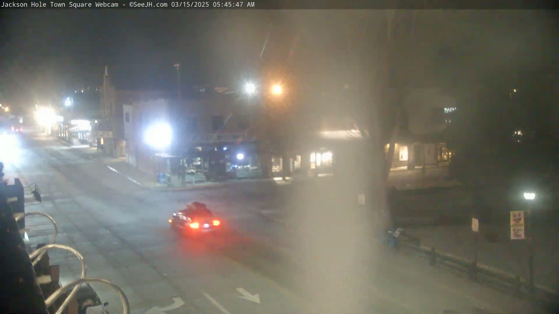 A foggy night scene shows a mostly empty street with a single car driving through a town square.