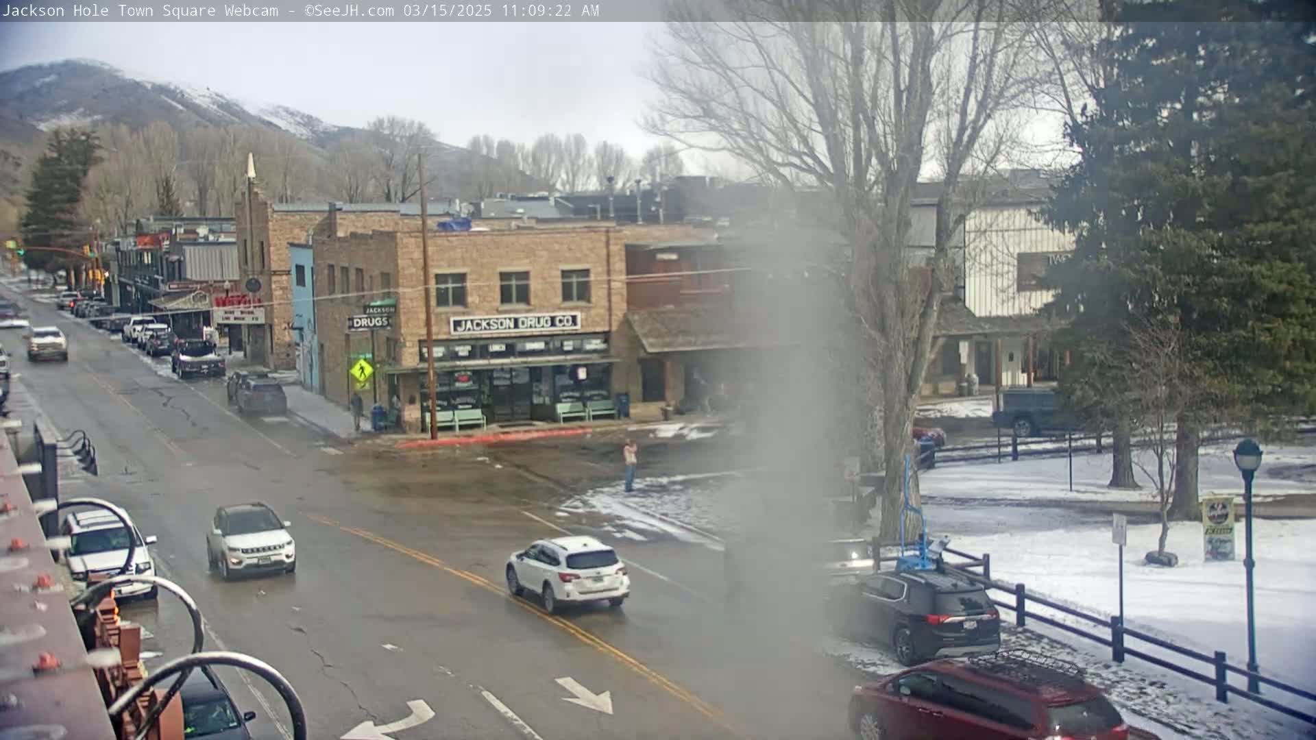 A town square scene on an overcast day shows several cars on wet streets, buildings with businesses, and a few pedestrians in front of a snowy park.