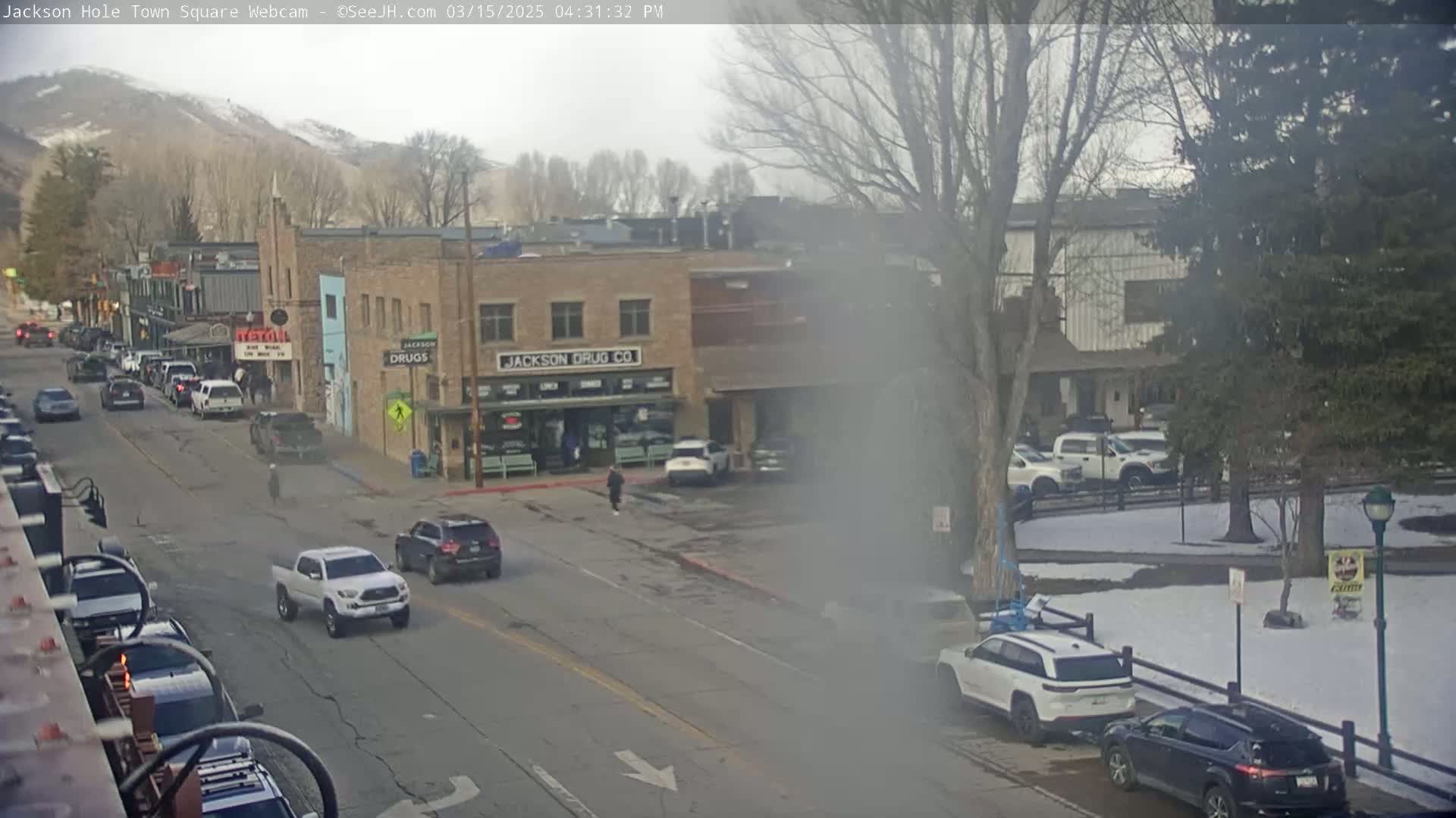 A hazy, overcast day reveals a town square with several vehicles parked and driving on a street lined with buildings, one of which is labeled "Jackson Drug Co."  There are bare trees and patches of snow visible.