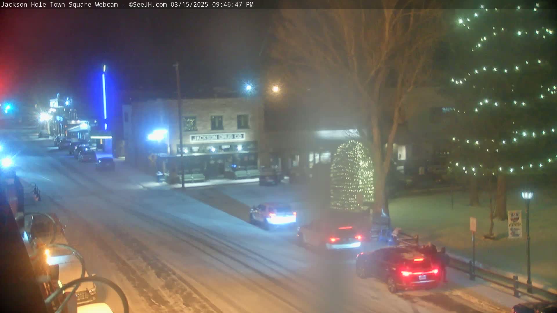 A snow-covered town square at night shows several cars driving on a street past buildings and a tree illuminated with Christmas lights.