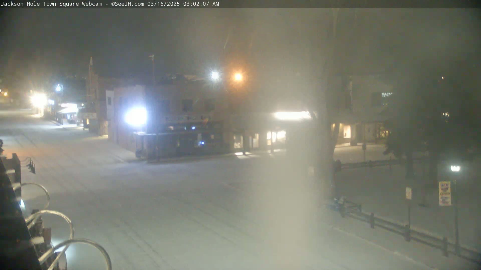 A snow-covered town square at night, illuminated by streetlights and building lights, with light snowfall obscuring visibility.