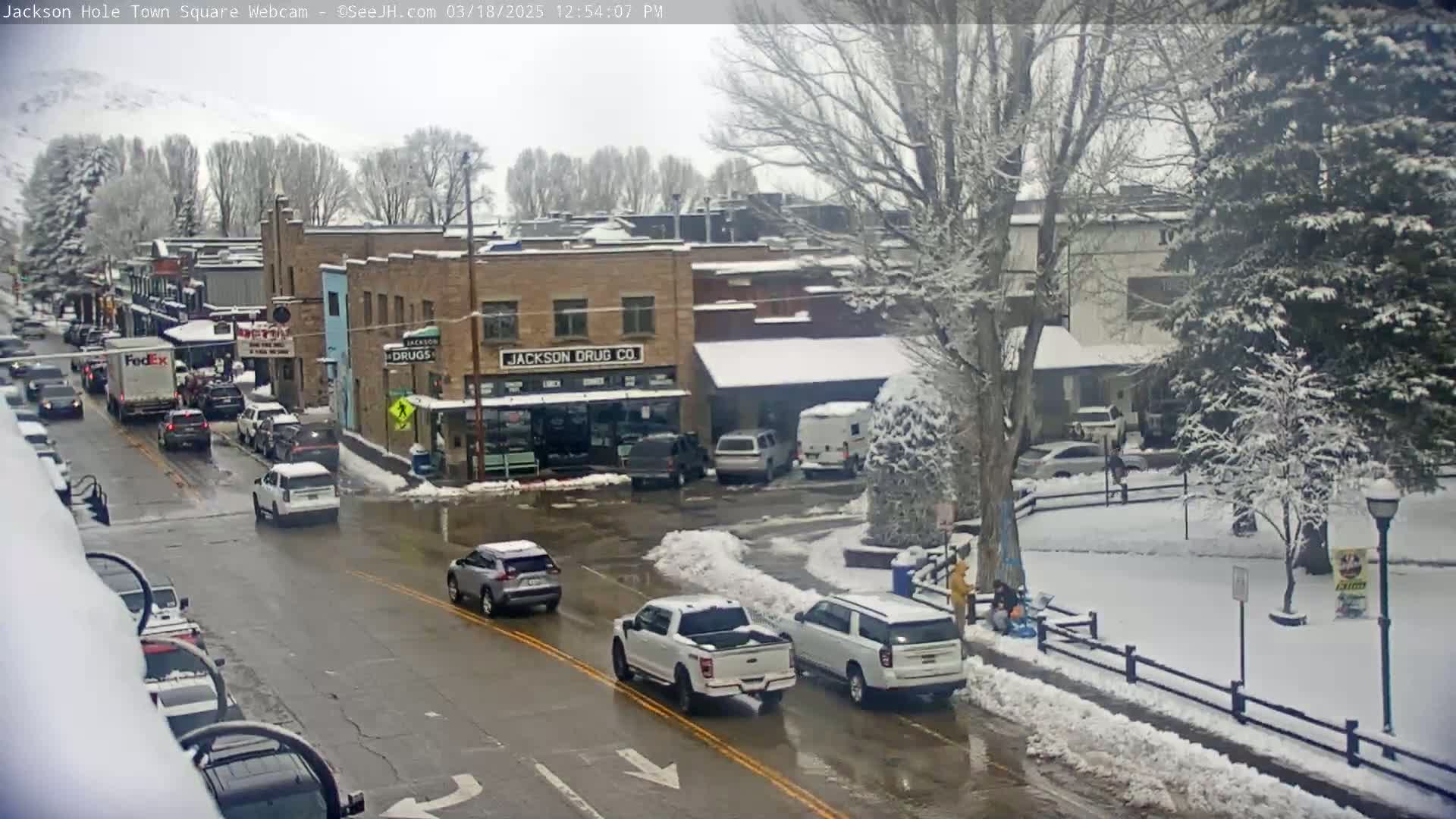 A snow-covered town square scene shows cars driving on a street past buildings, with snow-covered trees and a park in the background.