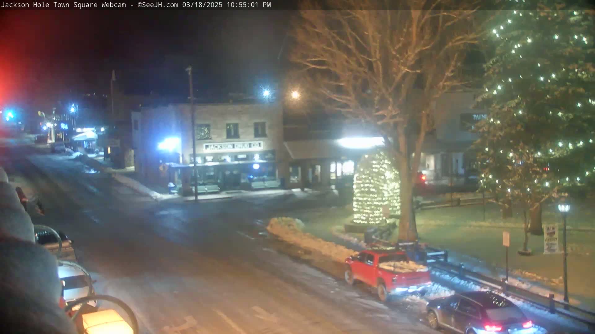 A nighttime view of a snow-covered town square with a few cars and a large illuminated tree.