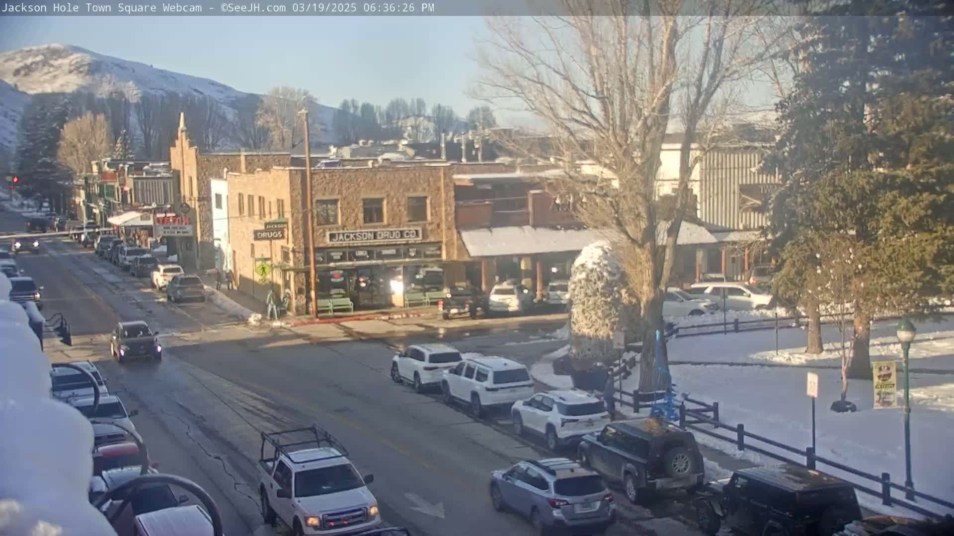 A sunny, snow-covered town square scene shows cars parked and driving along a street lined with low buildings, some with snow on their roofs.