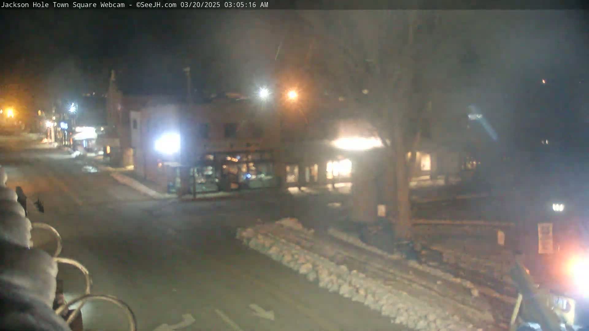 A nighttime view of a snow-covered town square, with buildings lit by streetlights and some snow piles visible along the road.