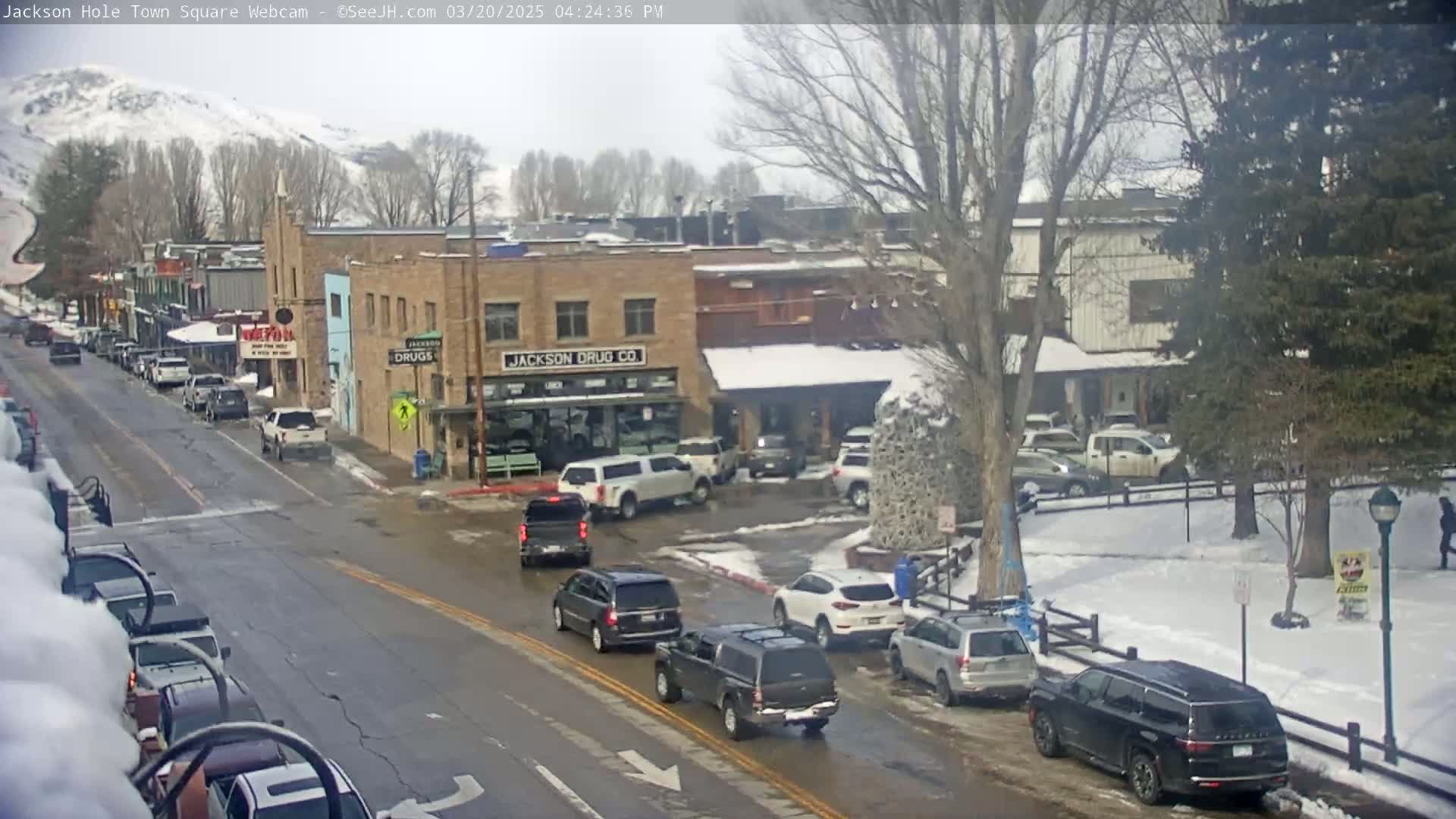 A street scene in a town shows several cars parked and driving on a wet road, with snow on the ground and in the background, under an overcast sky.