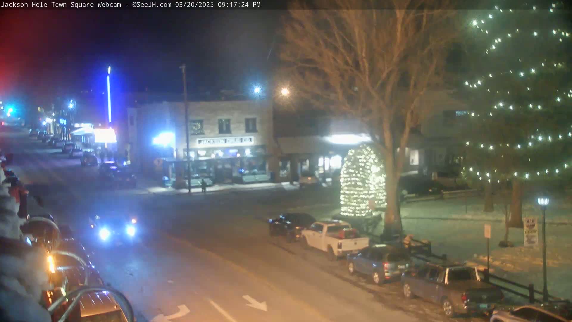 A nighttime view of a town square with cars parked along the street, a lit-up Christmas tree, and snow on the ground.