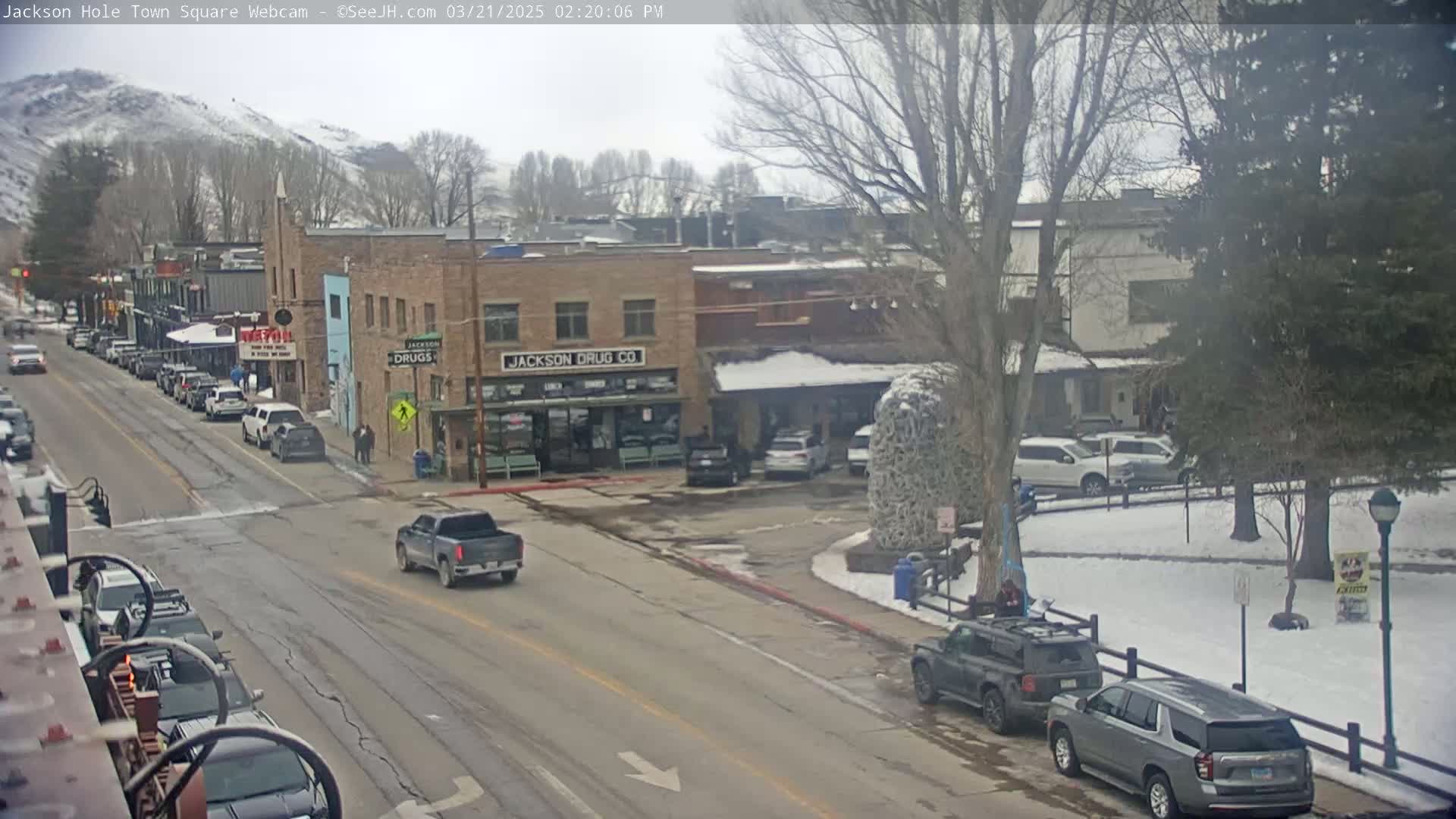 A town square with several cars parked and driving on a street, partially covered in snow, with a snow-covered hill in the background under an overcast sky.