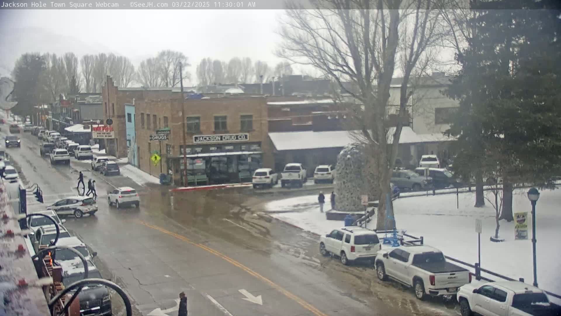 A snow-dusted town square with cars parked along the streets and pedestrians crossing, under an overcast sky.