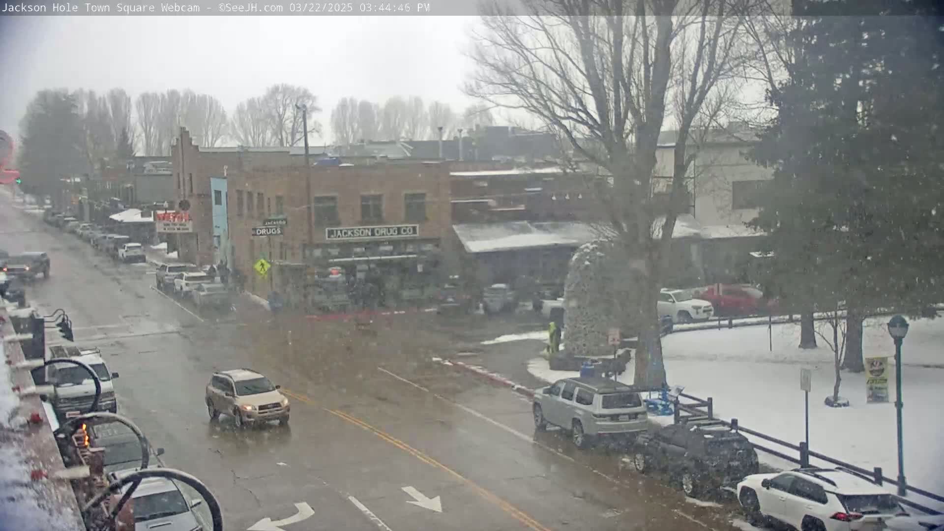 A snow-covered town square with cars driving on a wet street, several parked cars, and buildings in the background under a light snowfall.