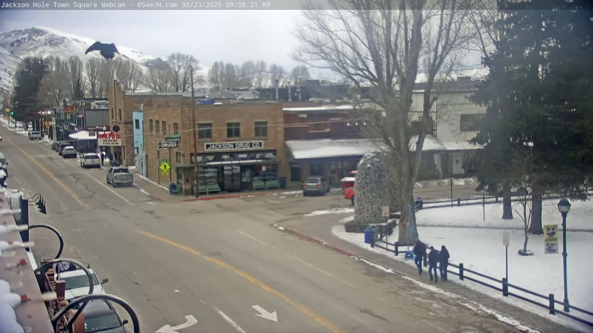 A town square with several buildings, cars parked along the street, and a few pedestrians walking on a snowy day.