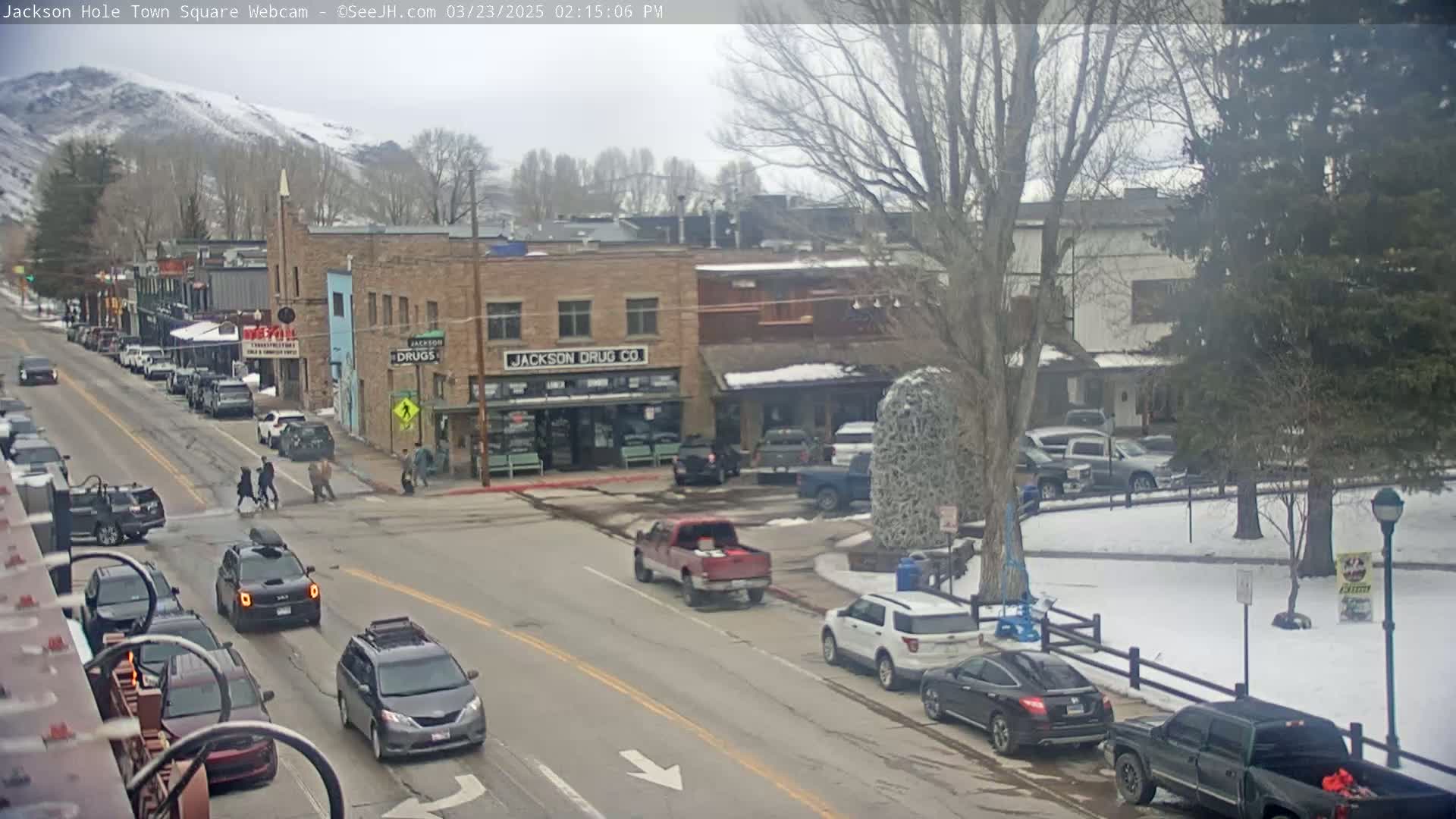 A town square with cars parked along the street and several pedestrians crossing a street intersection under an overcast sky with some snow on the ground.
