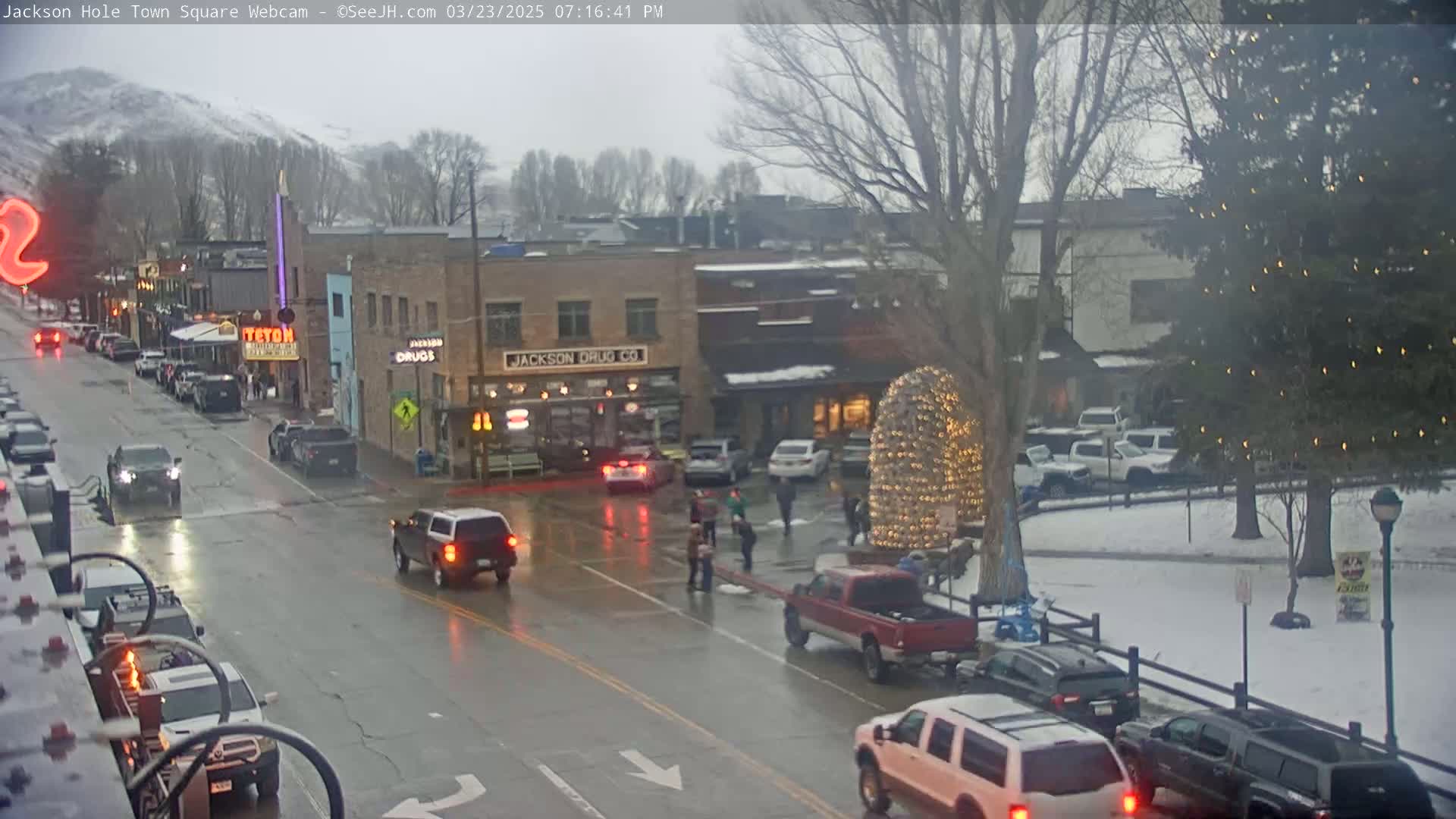 A wet town square with several vehicles driving on the street, people walking near a decorated tree, and snow on the ground in the background.