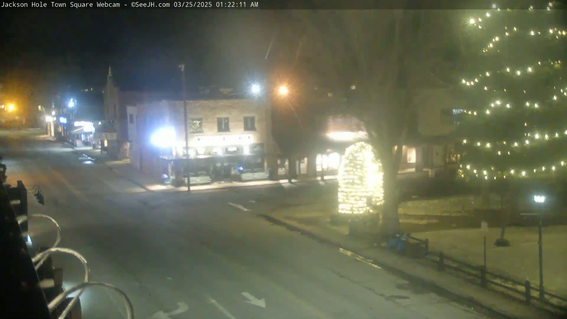A nighttime view of a town square with snow on the ground, illuminated by streetlights and a large, brightly lit Christmas tree.