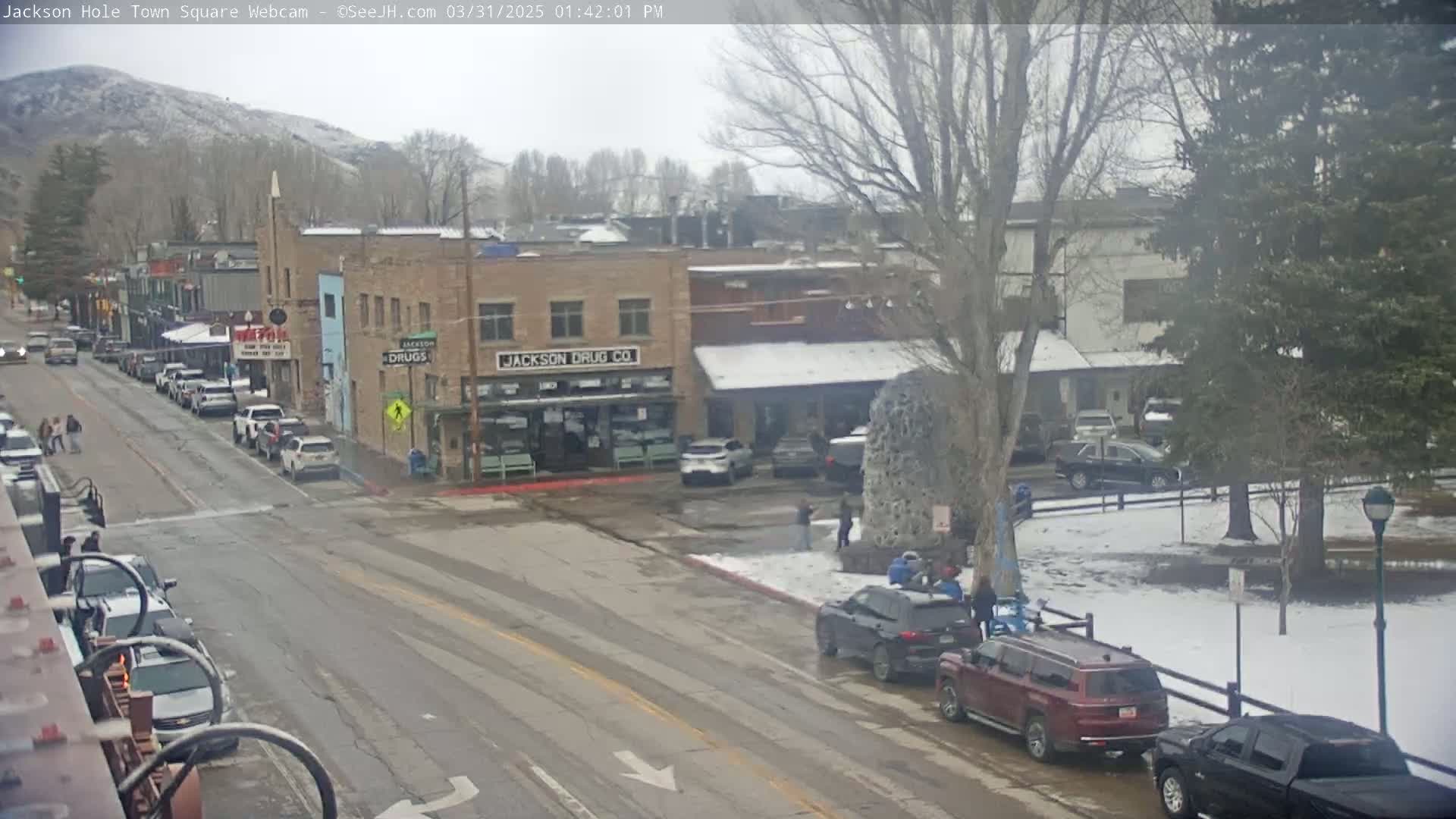 A snow-dusted town square with parked cars and pedestrians, under an overcast sky.