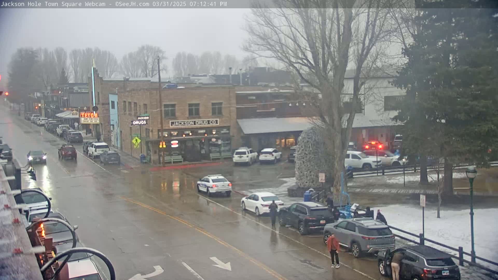 A rain-slicked town square scene shows several cars, some police vehicles, and pedestrians near buildings, one of which is a drug store.