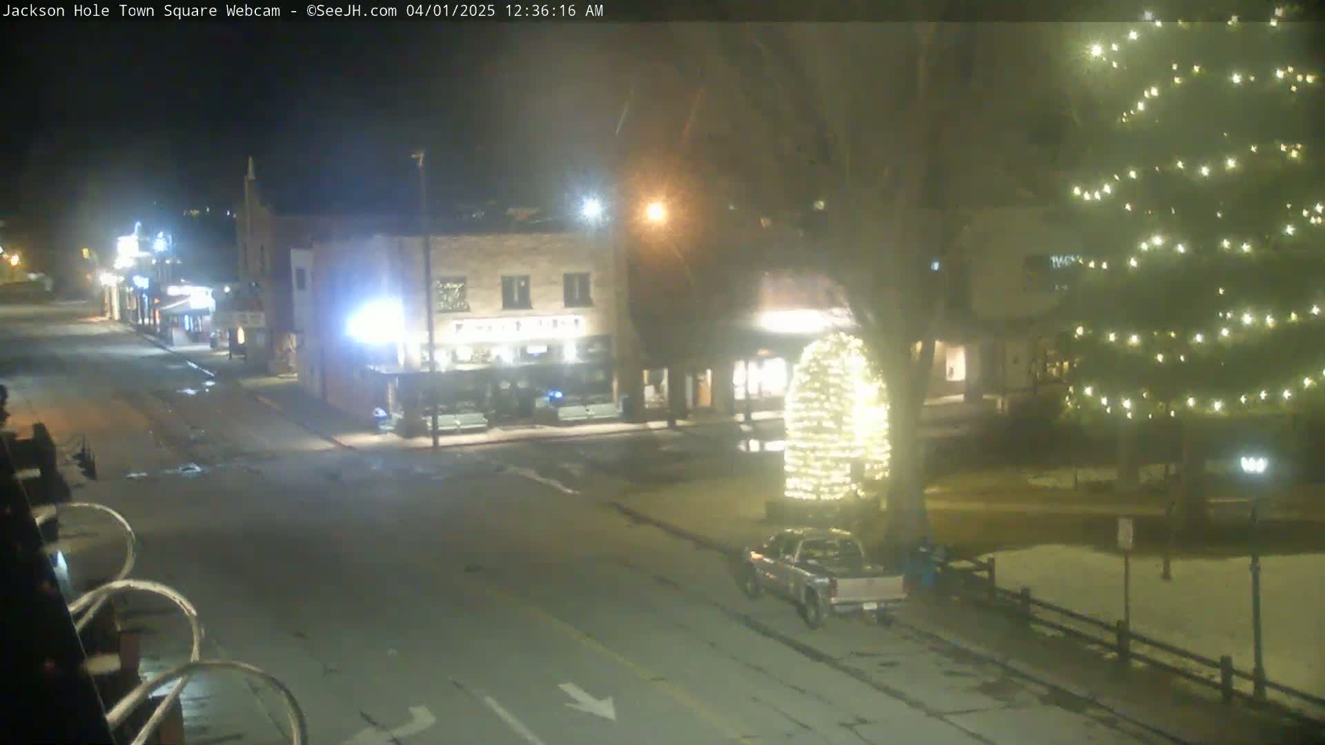 A nighttime view of a town square with a few buildings, a street, a pickup truck, and a large evergreen tree decorated with lights, under lightly snowy conditions.