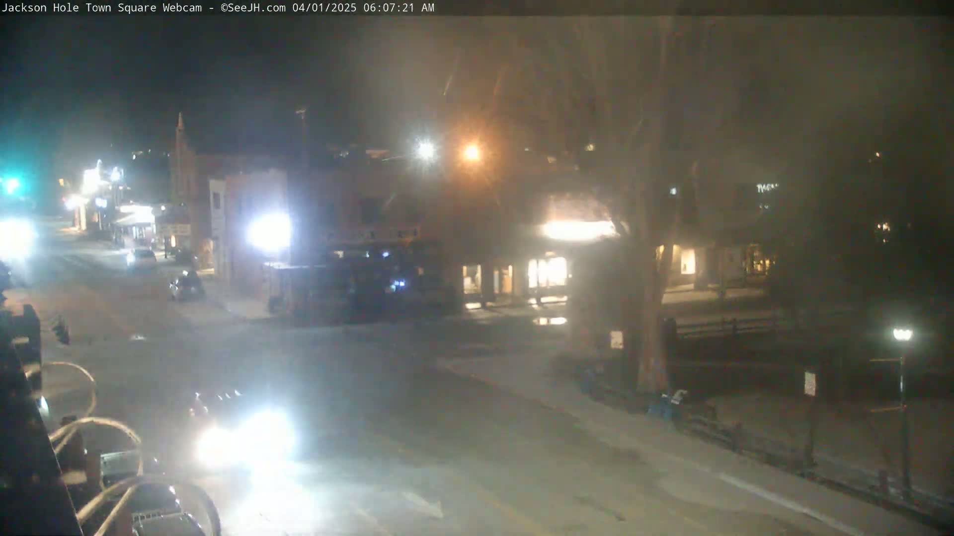 A nighttime, snow-covered town square shows a few vehicles on the road and buildings with lights on.