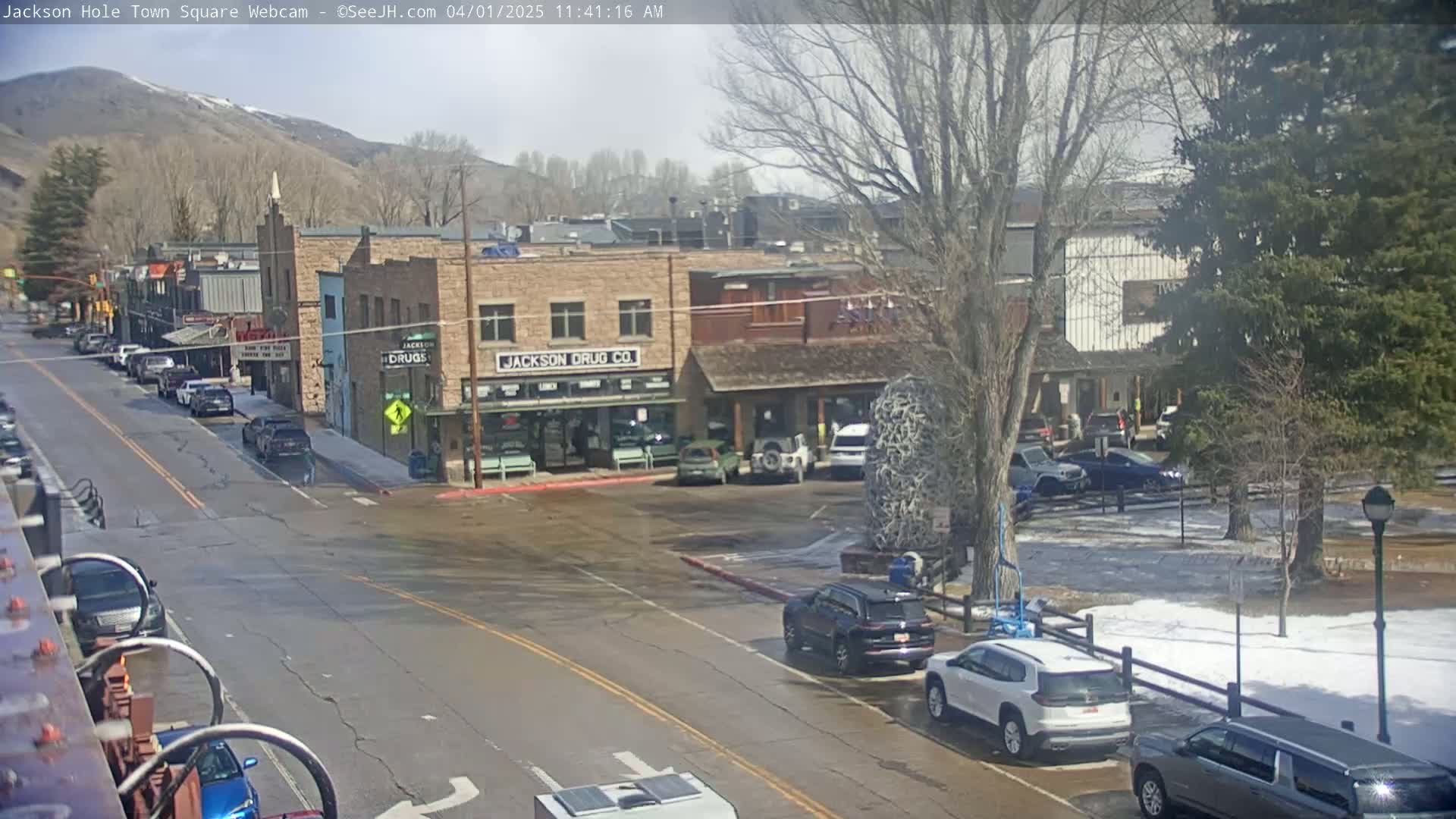 A partly sunny day reveals a town square with parked cars, buildings, and trees, some with patches of snow visible.
