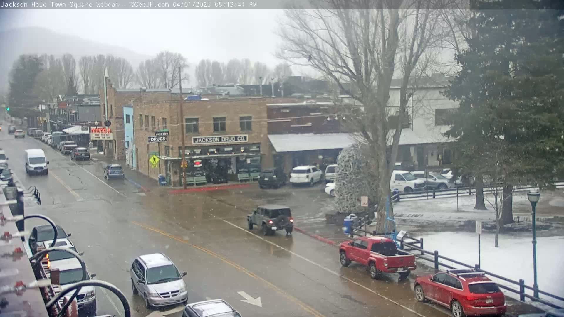 A town square, lightly snowing, with cars driving on wet streets and parked in a snowy lot, in front of brick buildings.