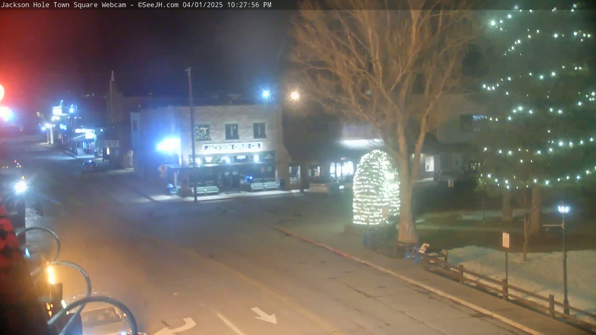 A nighttime view of a town square with a street, buildings, and trees decorated with lights, a little snow on the ground.
