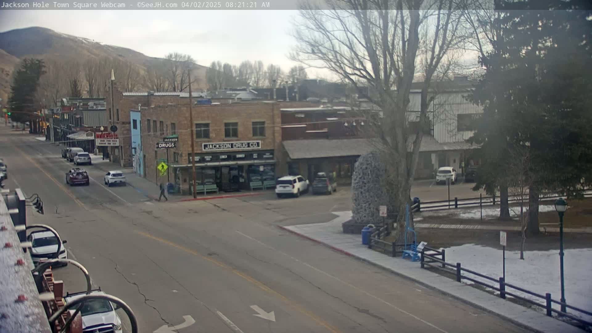 A mostly clear, cold day reveals a town square with several cars parked and driving on the street, buildings lining the street, and a small park with snow on the ground.