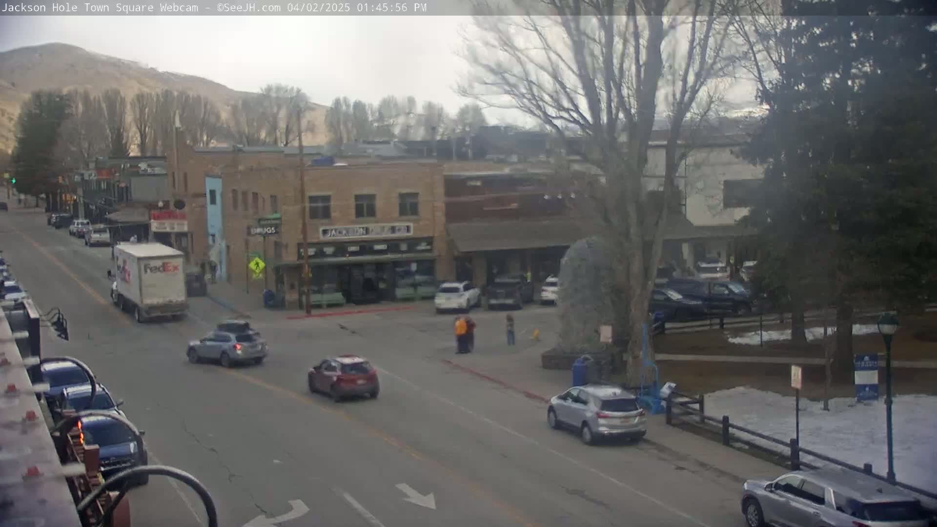 A town square with several cars and a FedEx truck on a street, under an overcast sky with some snow visible on the ground.