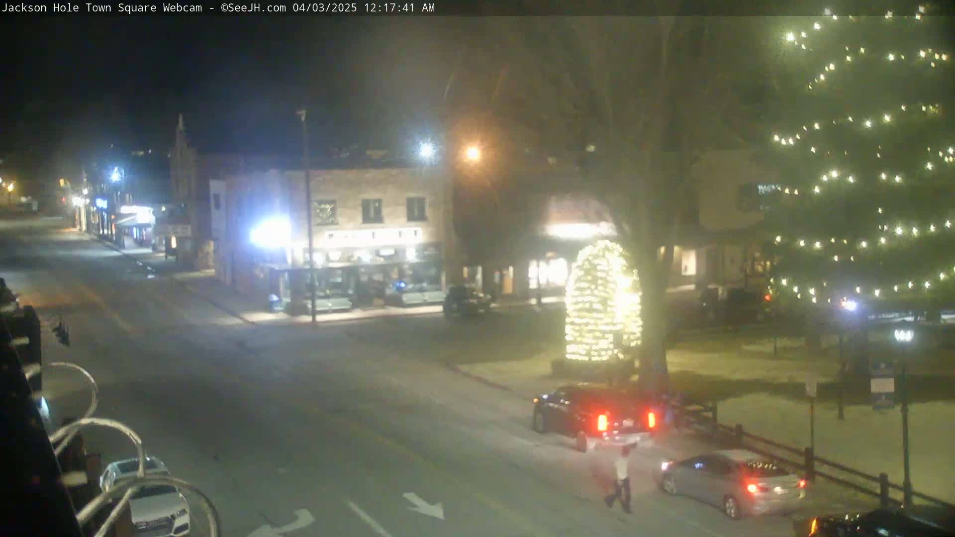 A nighttime view of a town square with a large illuminated Christmas tree, cars parked and driving on the road, and a pedestrian walking.