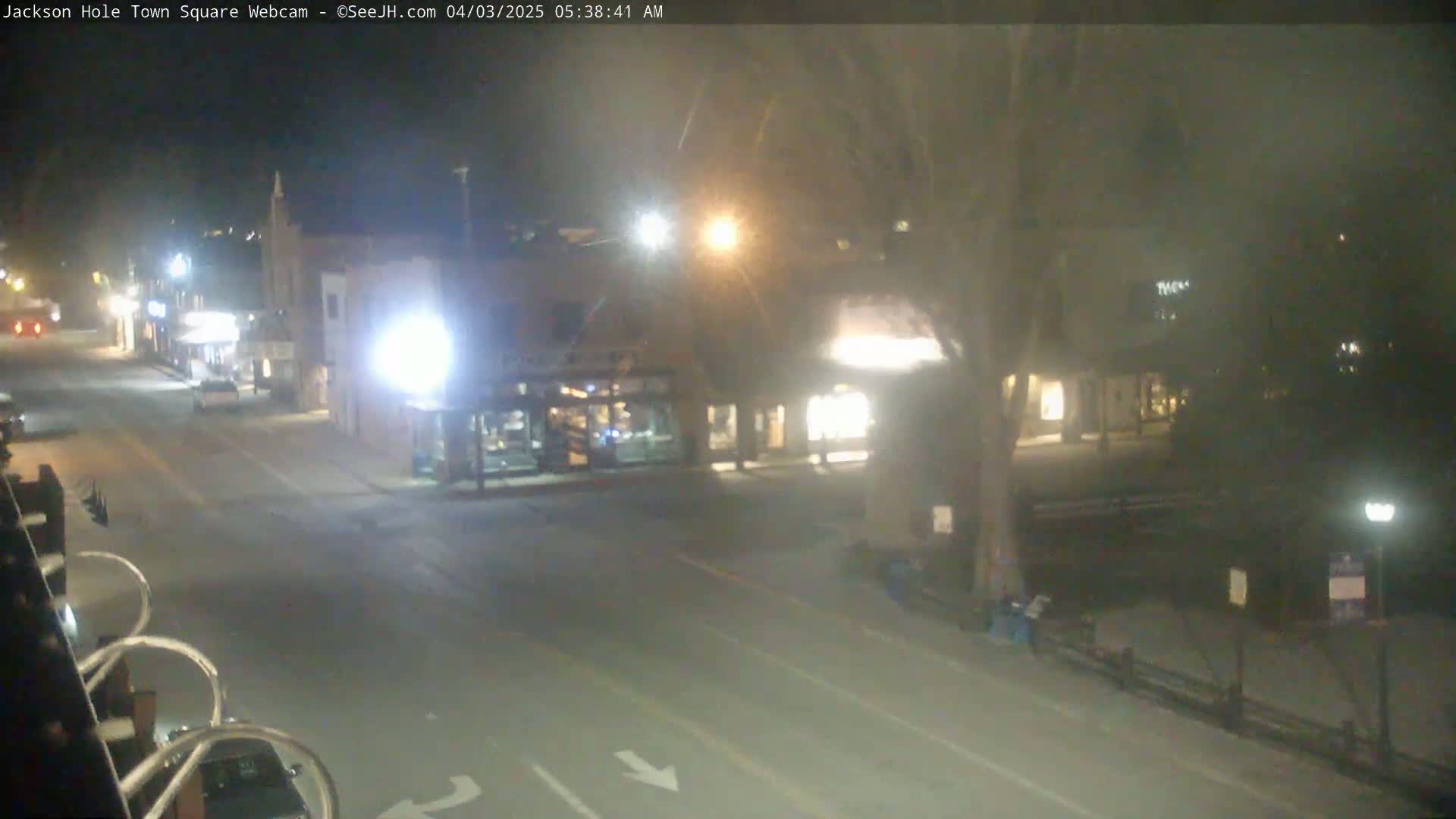 A nighttime view of a mostly empty town square with several buildings, streetlights, and a few cars, under hazy or foggy conditions.