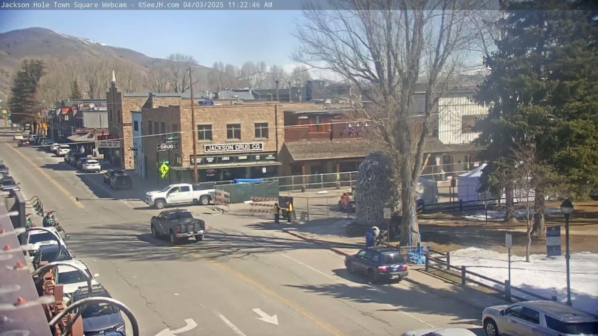 A sunny day shows a town square with several cars and trucks on the street, construction barriers, and some people walking near a large tree in a park-like setting, with mountains in the background.