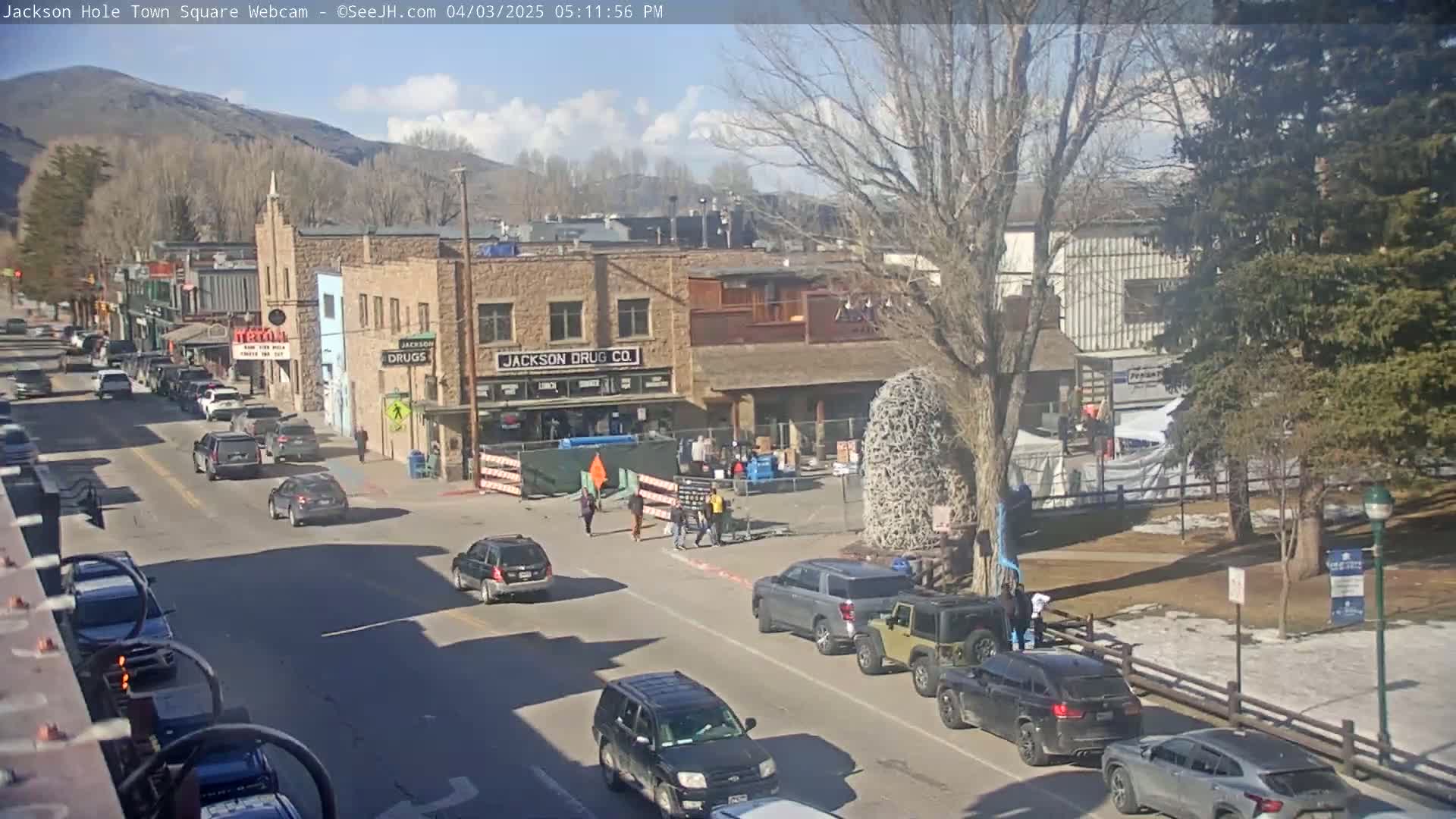 A sunny day in a town square with cars parked and driving on a street lined with buildings, some trees, and people walking.