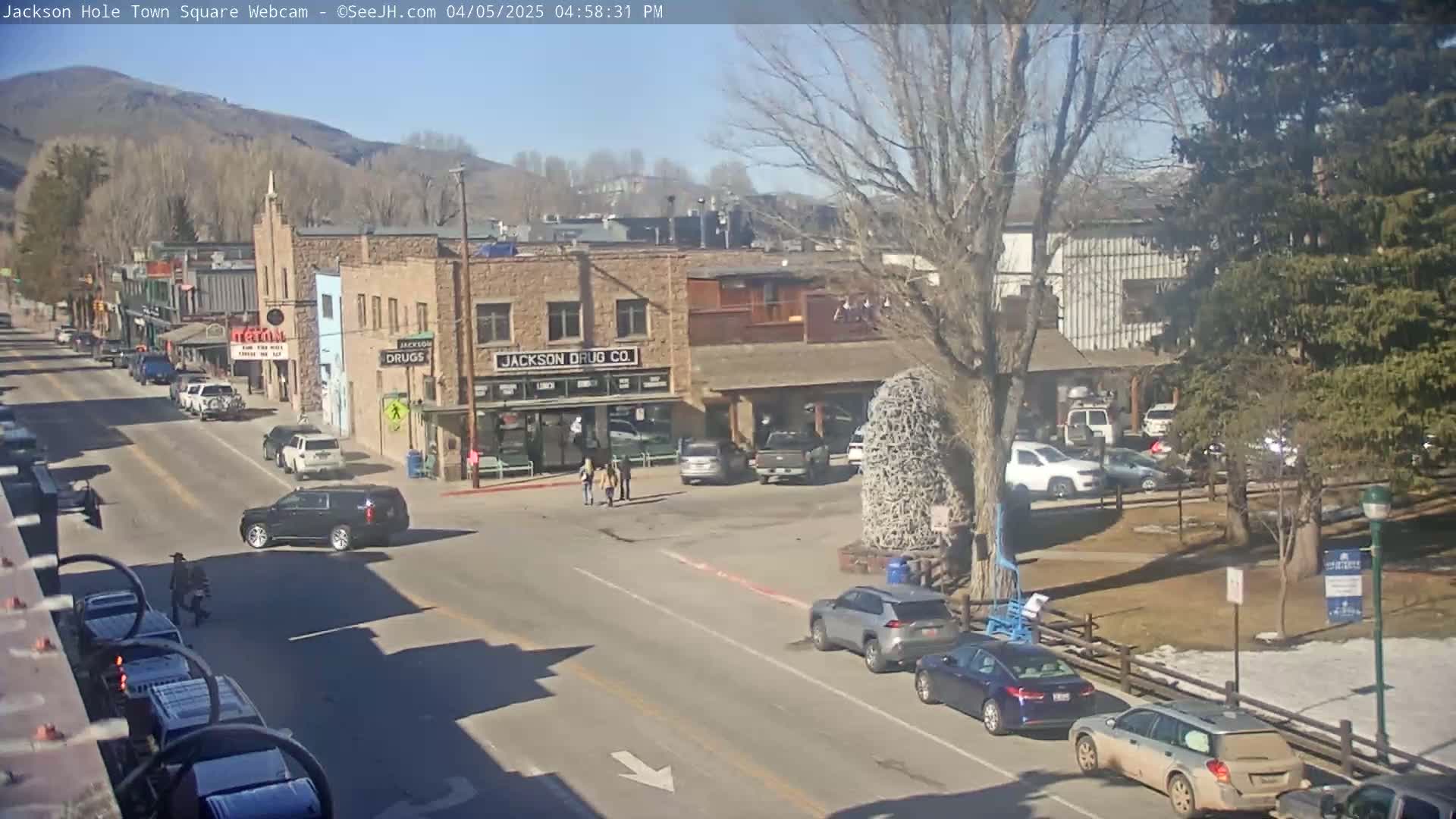 A sunny day in a town square, with several cars parked and driving on the street, some pedestrians walking near buildings, and trees with bare branches.